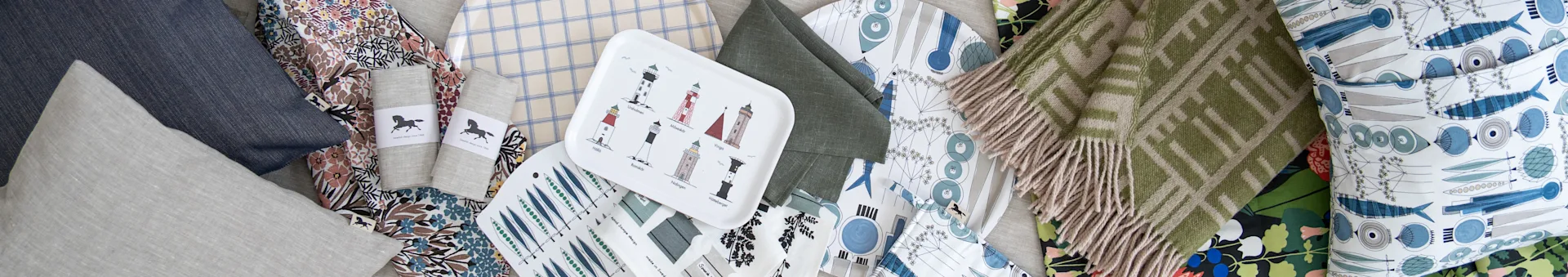 Assortment of home textiles: blue and grey pillows, floral and plaid fabrics, lighthouse tray, green patterned blanket.
