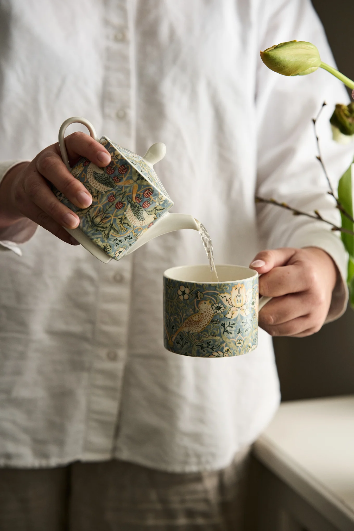 Hands pouring from a blue teapot with a bird and berry pattern into a matching mug.