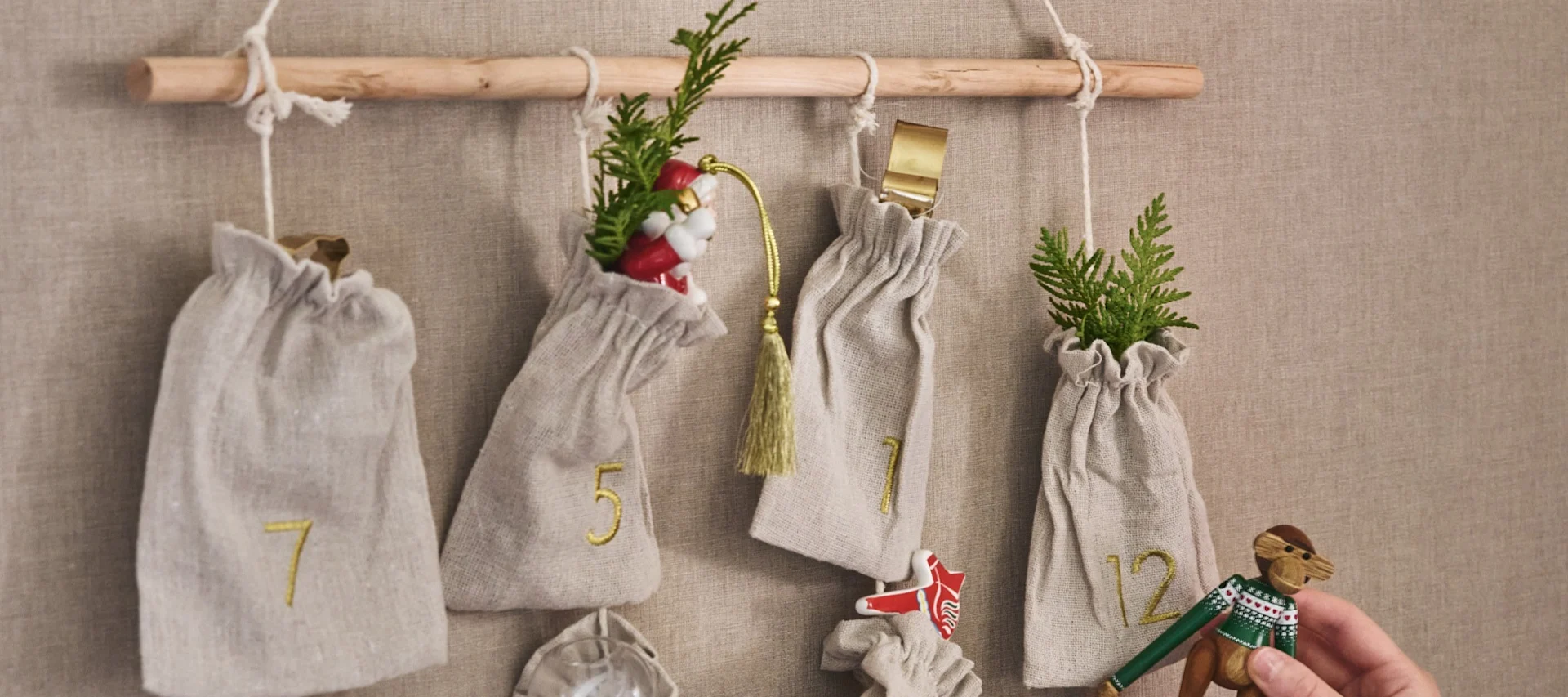 An advent calendar with small, numbered fabric bags hanging from a wooden rod. The bags contain festive items like evergreen sprigs, a Santa figure, and a wooden monkey toy.
