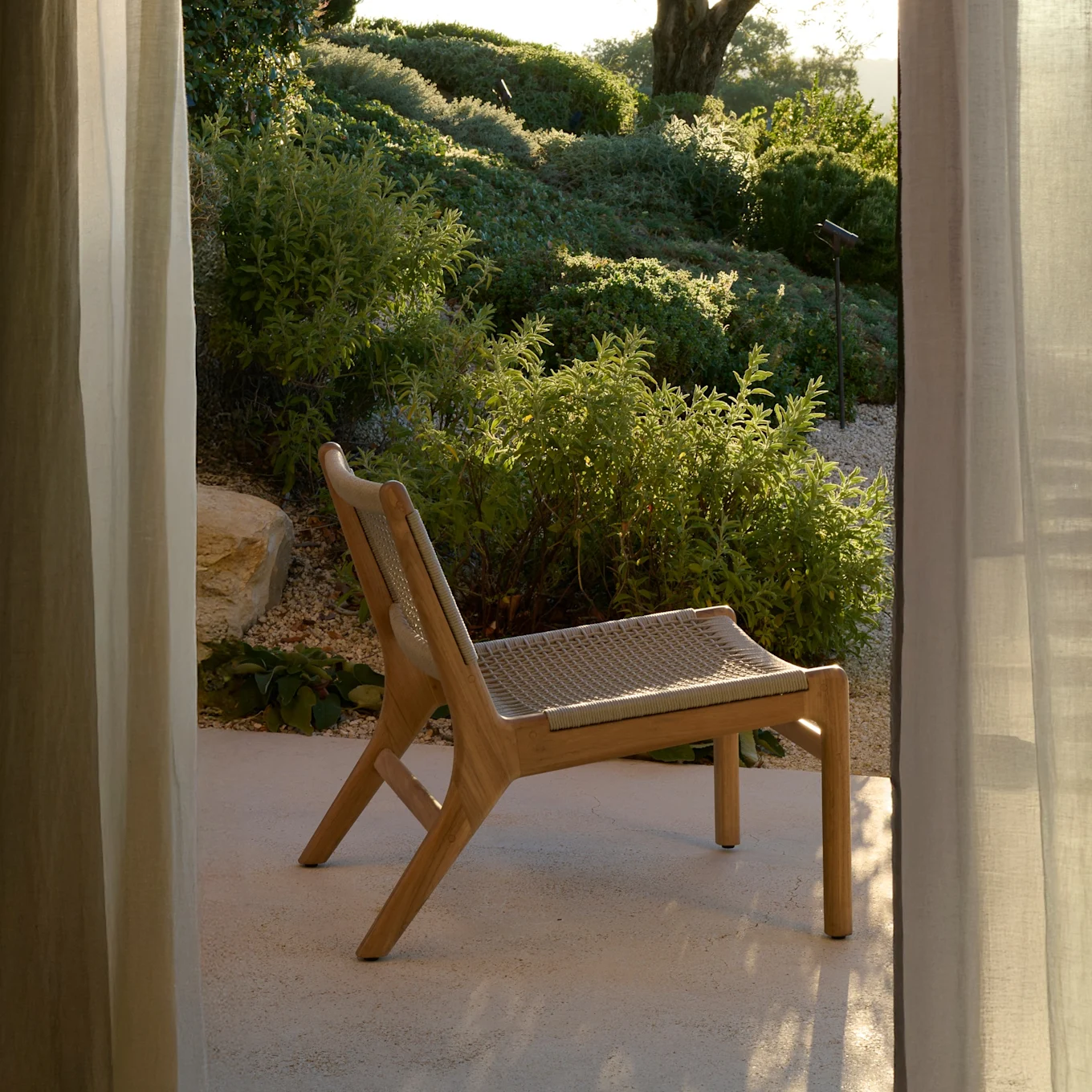 Modern wooden lounge chair with woven seat on a sunny patio, framed by curtains, overlooking a lush green garden.