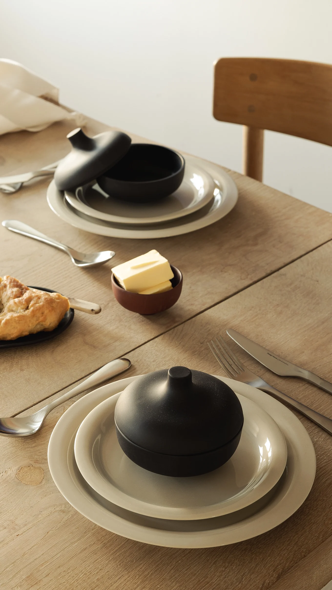 Table set for a meal with plates, black lidded bowls, silverware, butter, and bread on a wooden table.