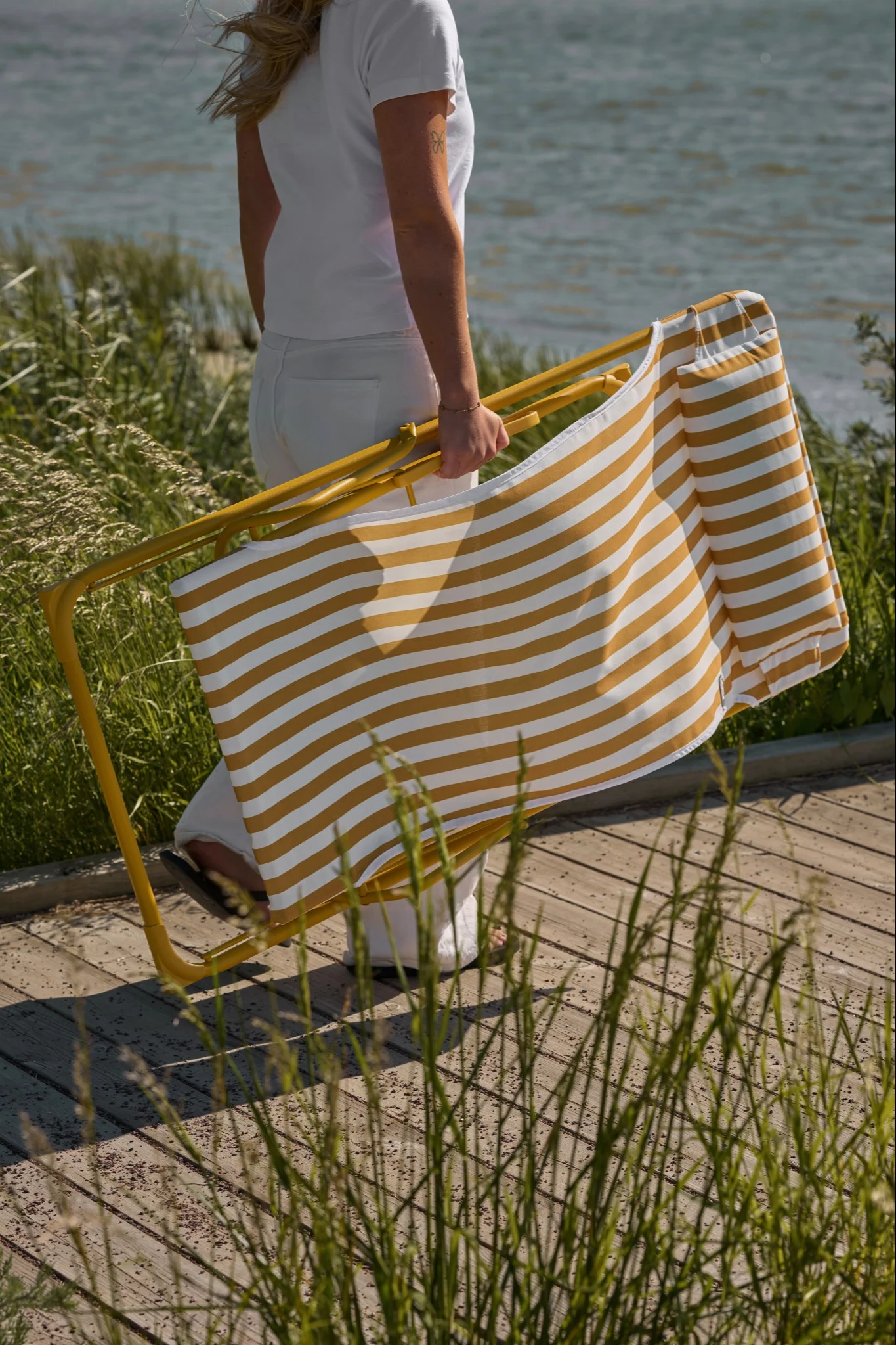 Person in white carries a folded yellow and white striped beach chair on a boardwalk with tall grass by the water.
