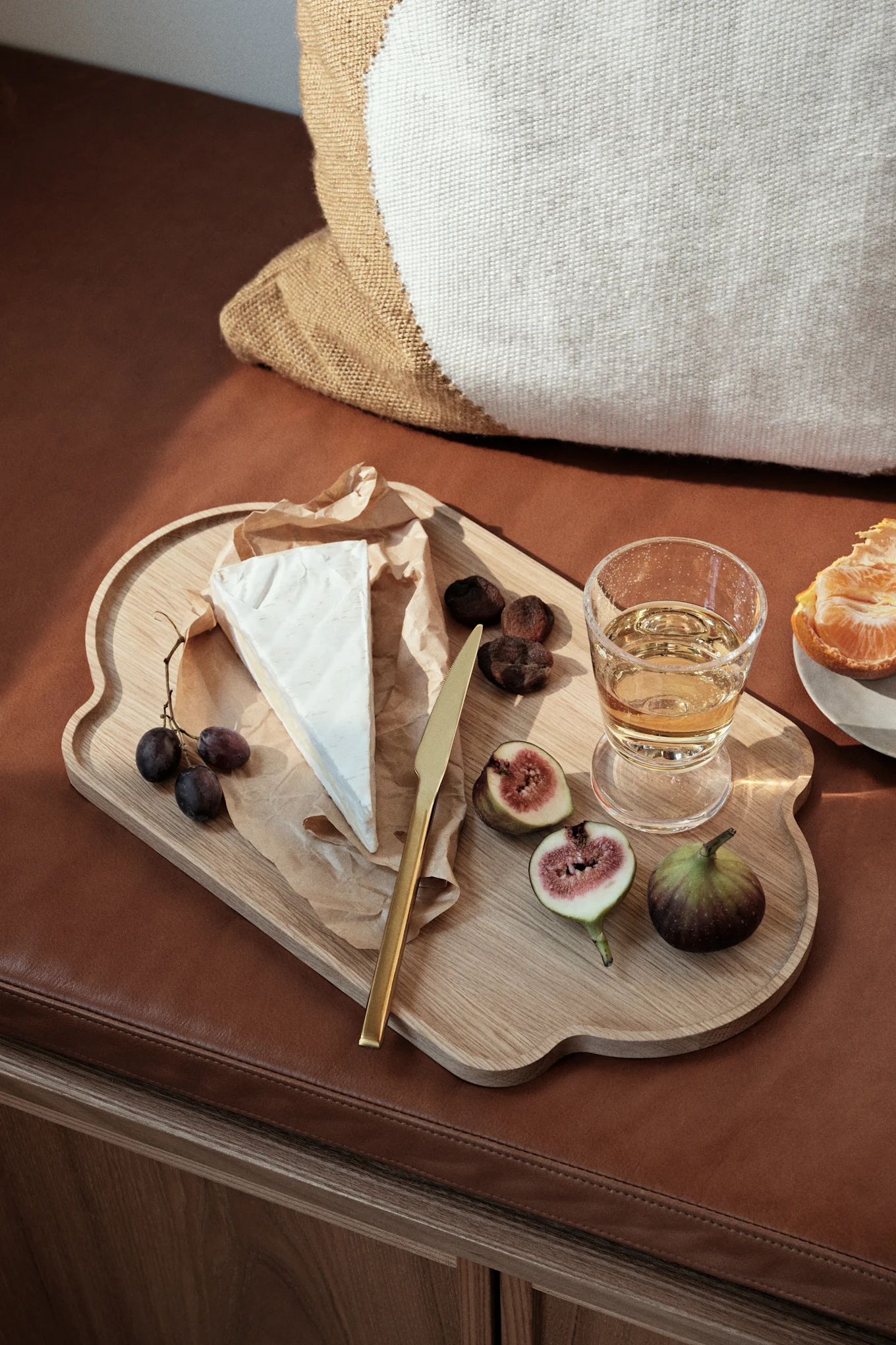 A wooden serving tray holds brie cheese, grapes, figs, dried apricots, a gold knife, and a glass of white wine on a brown leather surface.