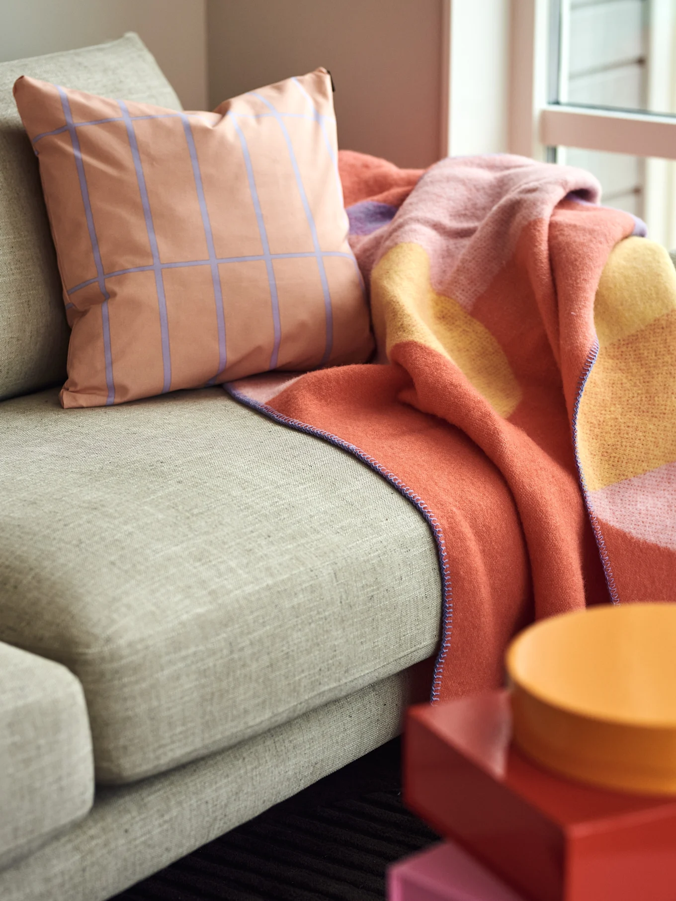 Peach grid-patterned pillow and colorful blanket on a beige sofa, with red and orange decor.