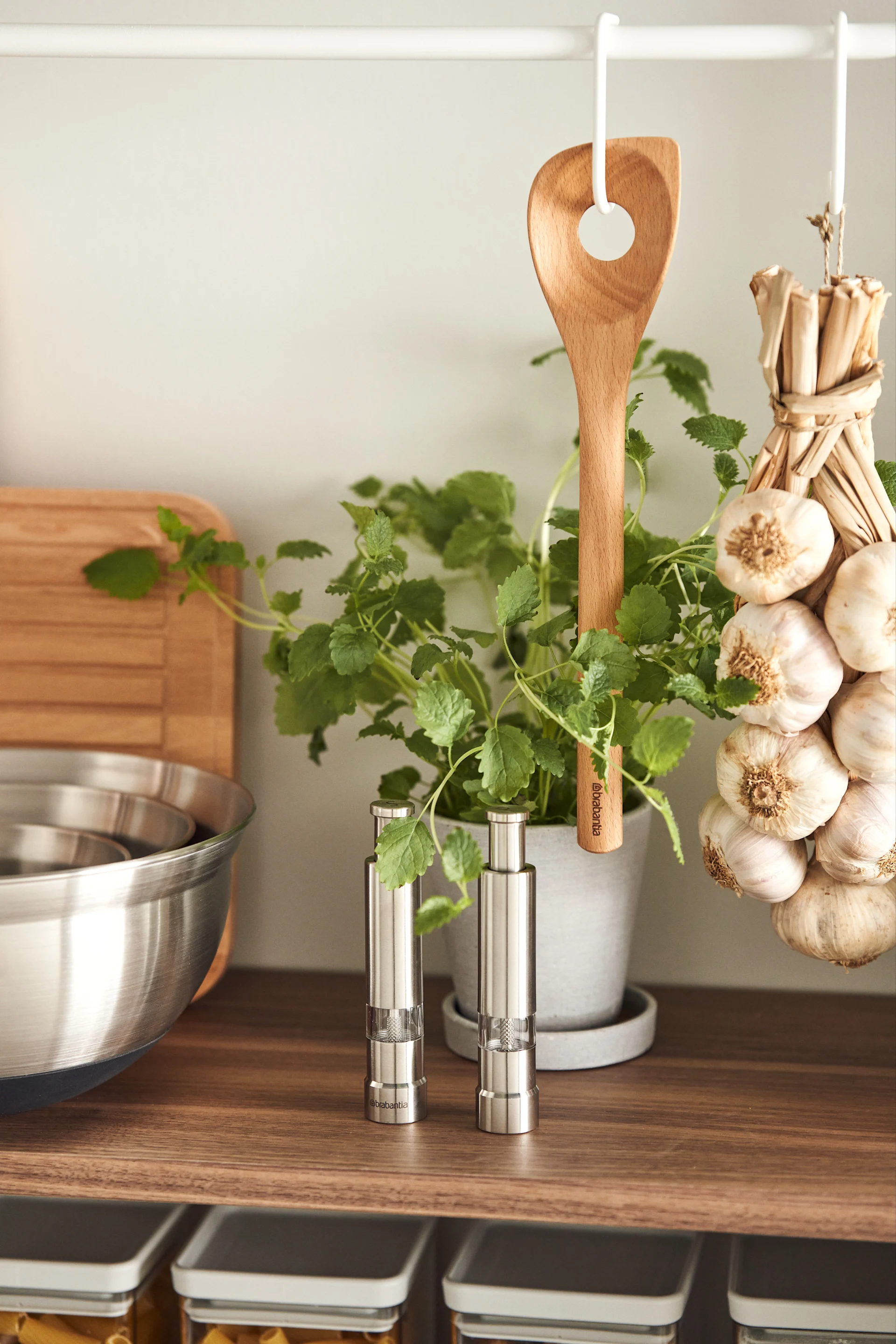 Kitchen shelf with stainless steel spice mills, a potted herb, wooden spoon, and hanging garlic.