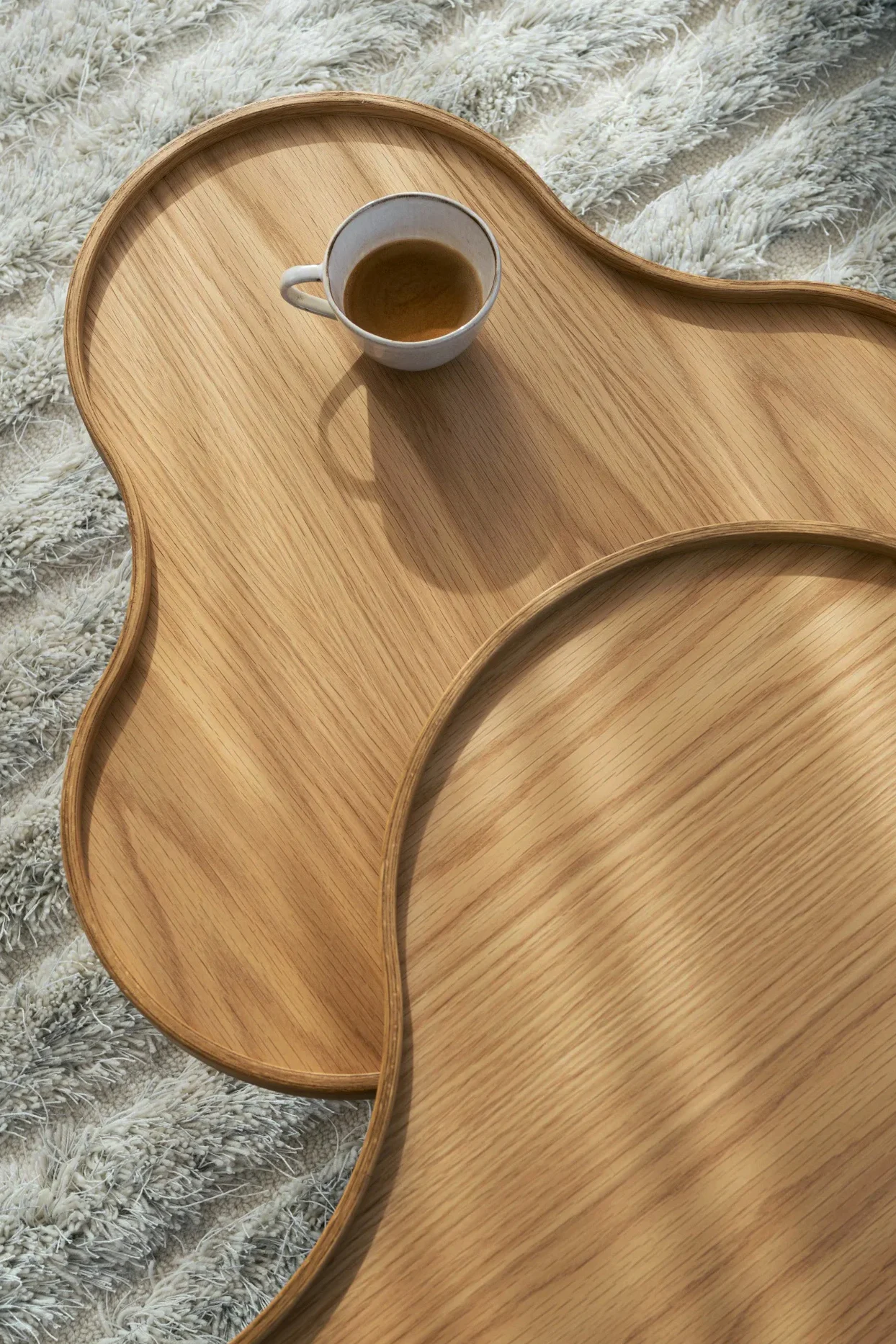 Overhead view of two overlapping light wooden trays with organic shapes on a white shaggy rug, one holding a cup of coffee.