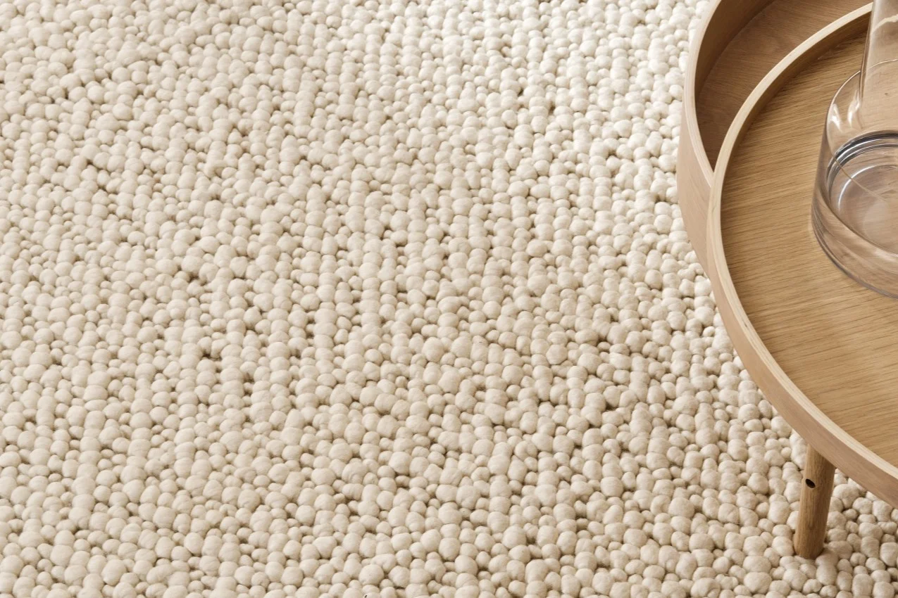 Close-up of a cream bobble rug with a light wood round side table and glass carafe.