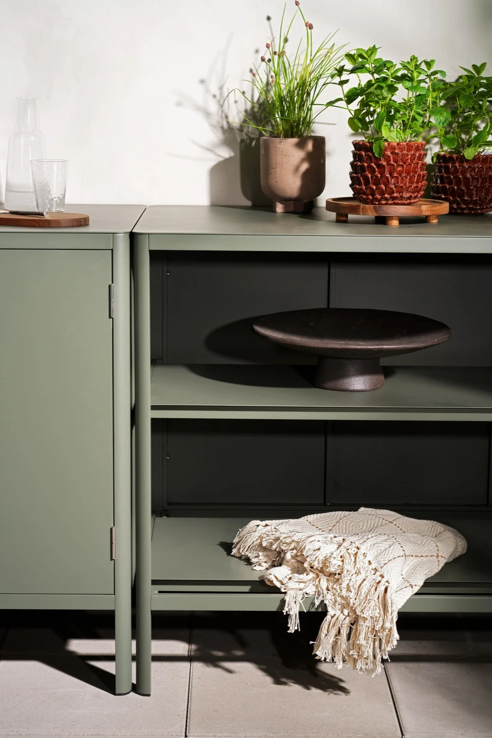 Sage green cabinet and shelving unit, featuring potted plants, a glass bottle, a decorative bowl, and a textured blanket.