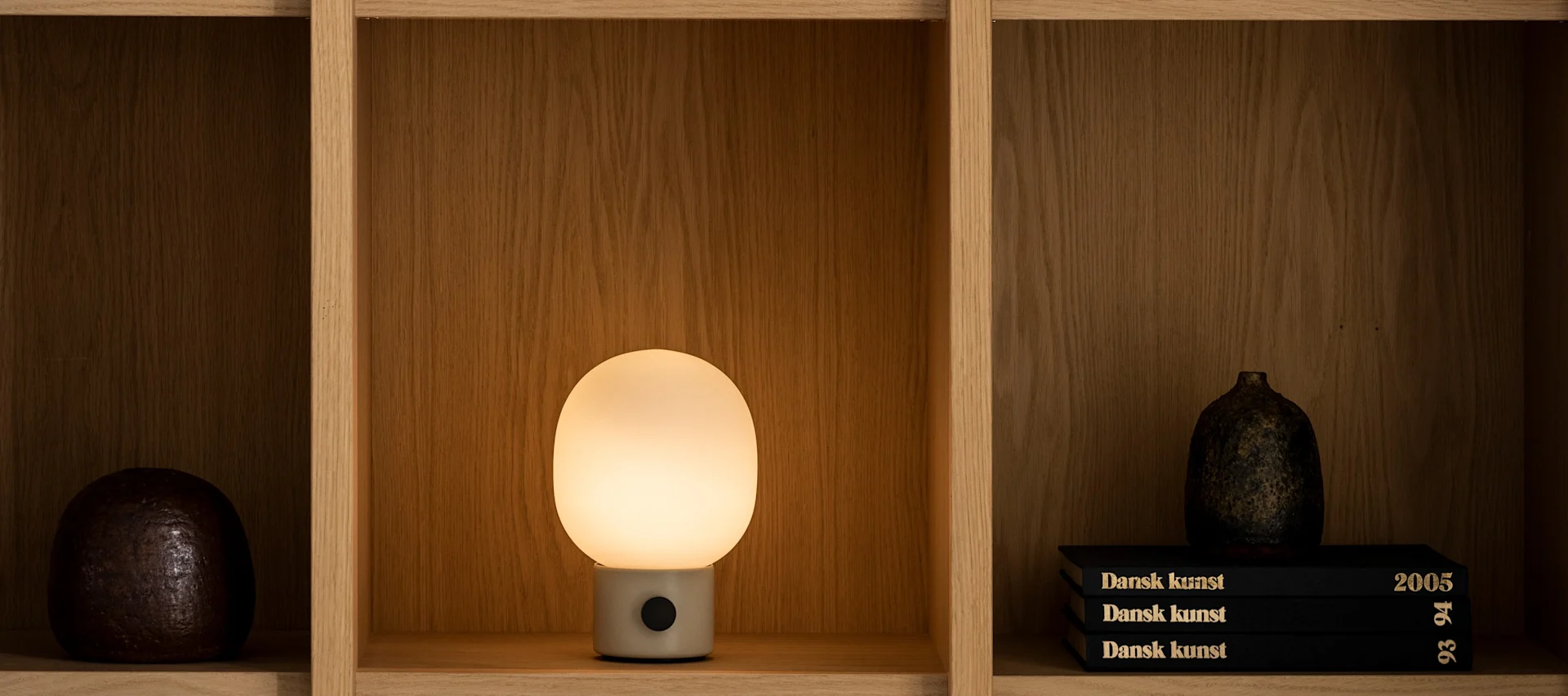 Wooden shelf displaying a dark spherical object, a glowing oval table lamp, and a stack of books with a dark vase.