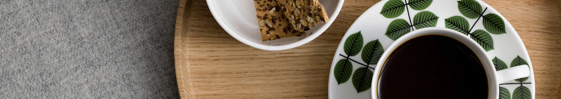 Coffee cup and saucer with green leaf pattern, next to a bowl of seed crackers on a wooden tray.