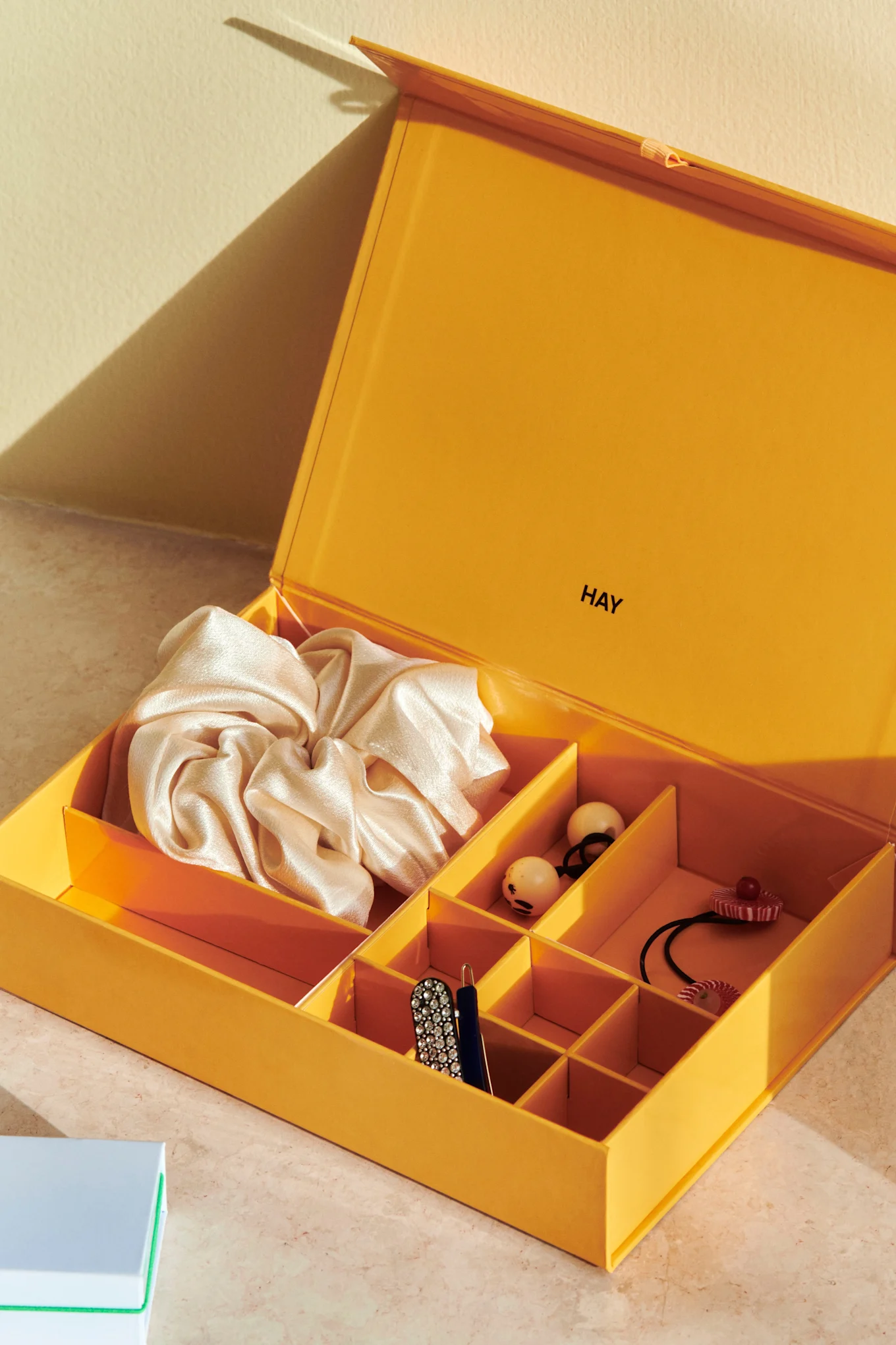 An open yellow storage box by HAY, containing a cream scrunchie, various hair accessories, a rhinestone hair clip, and a pen.