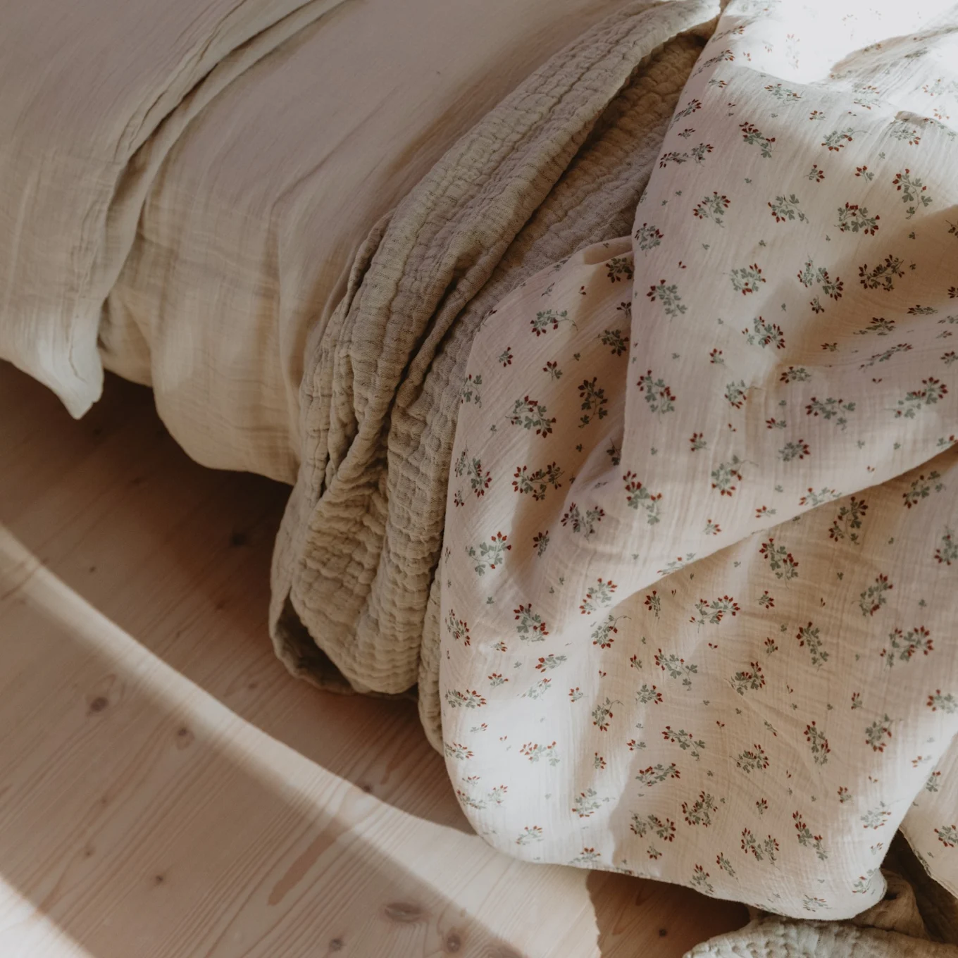 Light beige quilted bedding and a white floral duvet with red berries and green leaves on a bed with wooden floor.