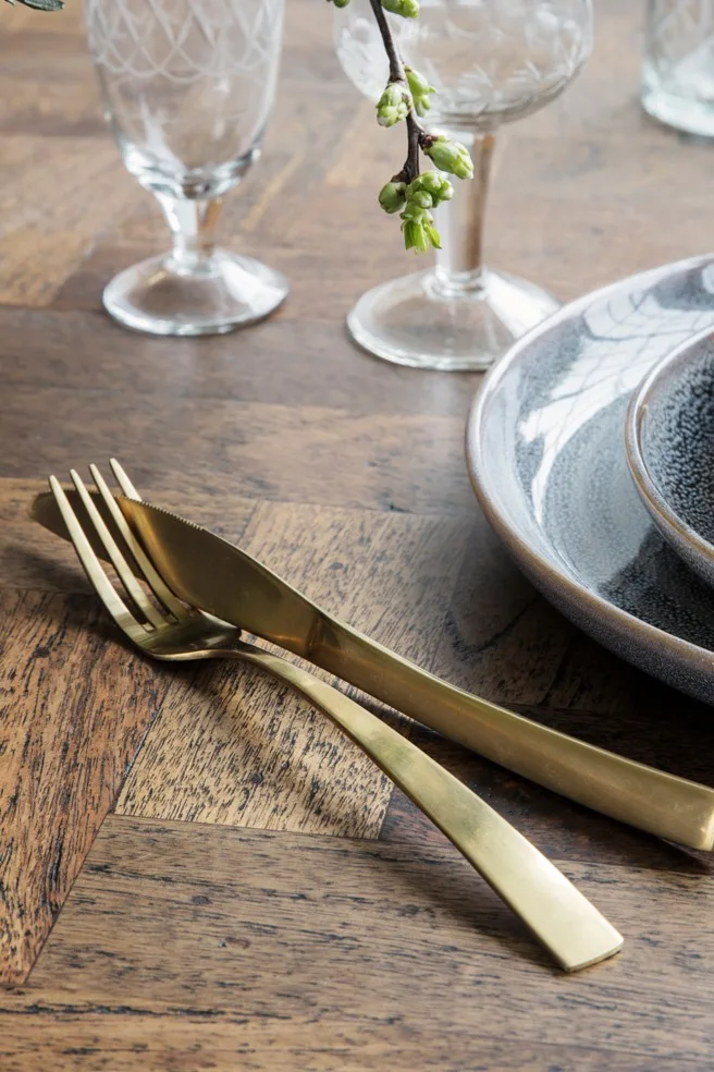 Gold fork and knife on a wooden table next to ceramic plates and patterned glasses, with a small branch of green buds.