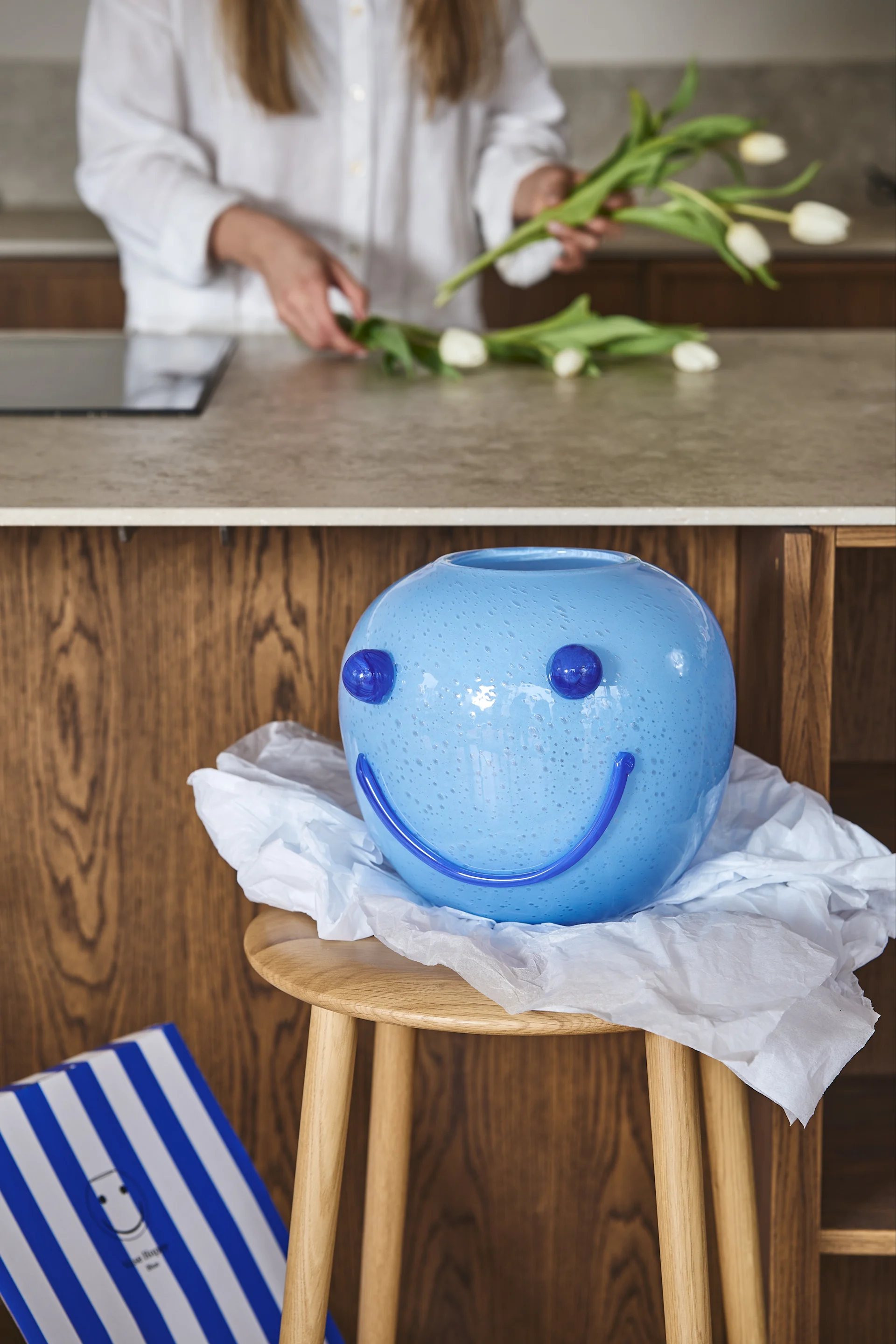 A light blue, round vase with a dark blue smiley face on a wooden stool, with a person arranging white tulips in the blurred background.