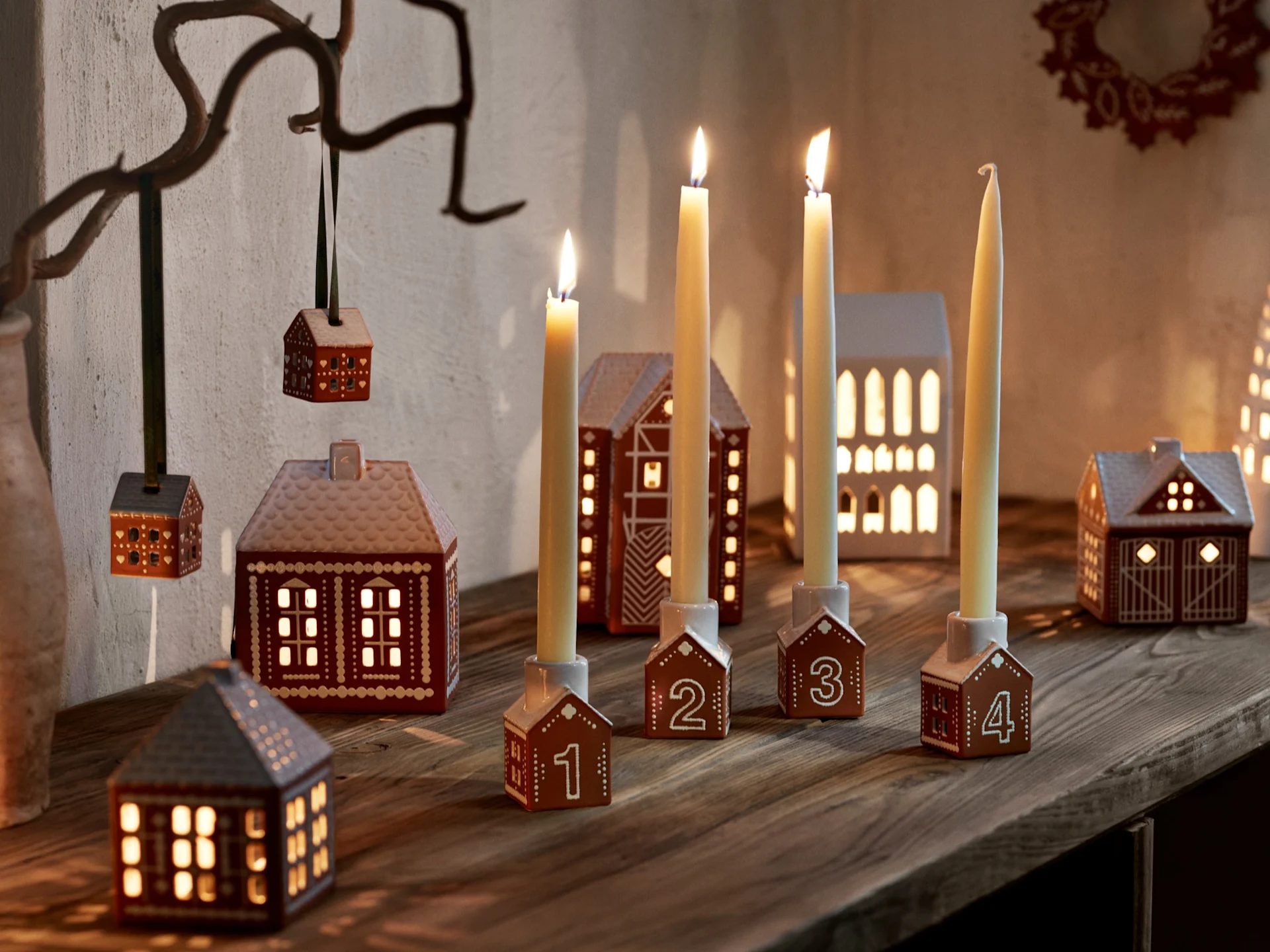 Four numbered gingerbread house candle holders with lit candles, and several gingerbread house lanterns on a wooden surface.