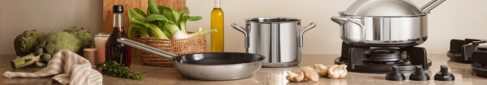 A kitchen counter with fresh vegetables, herbs, olive oil, and various stainless steel pots and pans, including a gas cooktop.