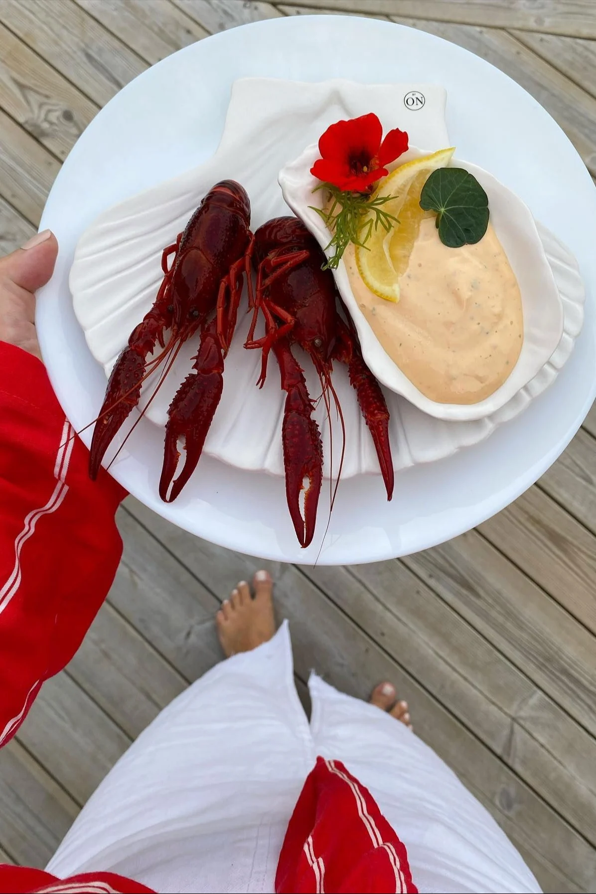 Overhead view of a person holding a plate with two red crawfish, a creamy sauce, lemon, and a red flower, on a wooden deck.
