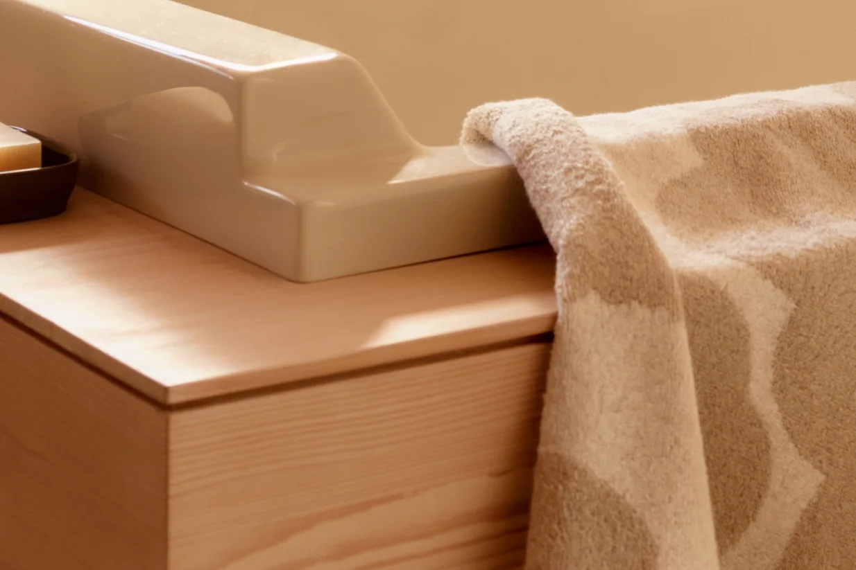 Close-up of a beige textured towel draped over a light-colored ceramic fixture on a natural wood vanity with a bar of soap.