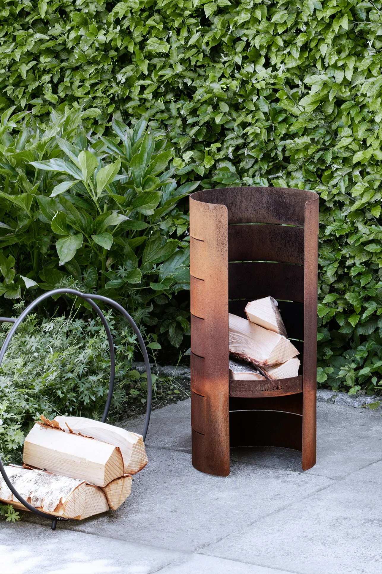 A rusty metal outdoor log store filled with firewood, next to a circular log holder on a patio, with a green hedge in the background.