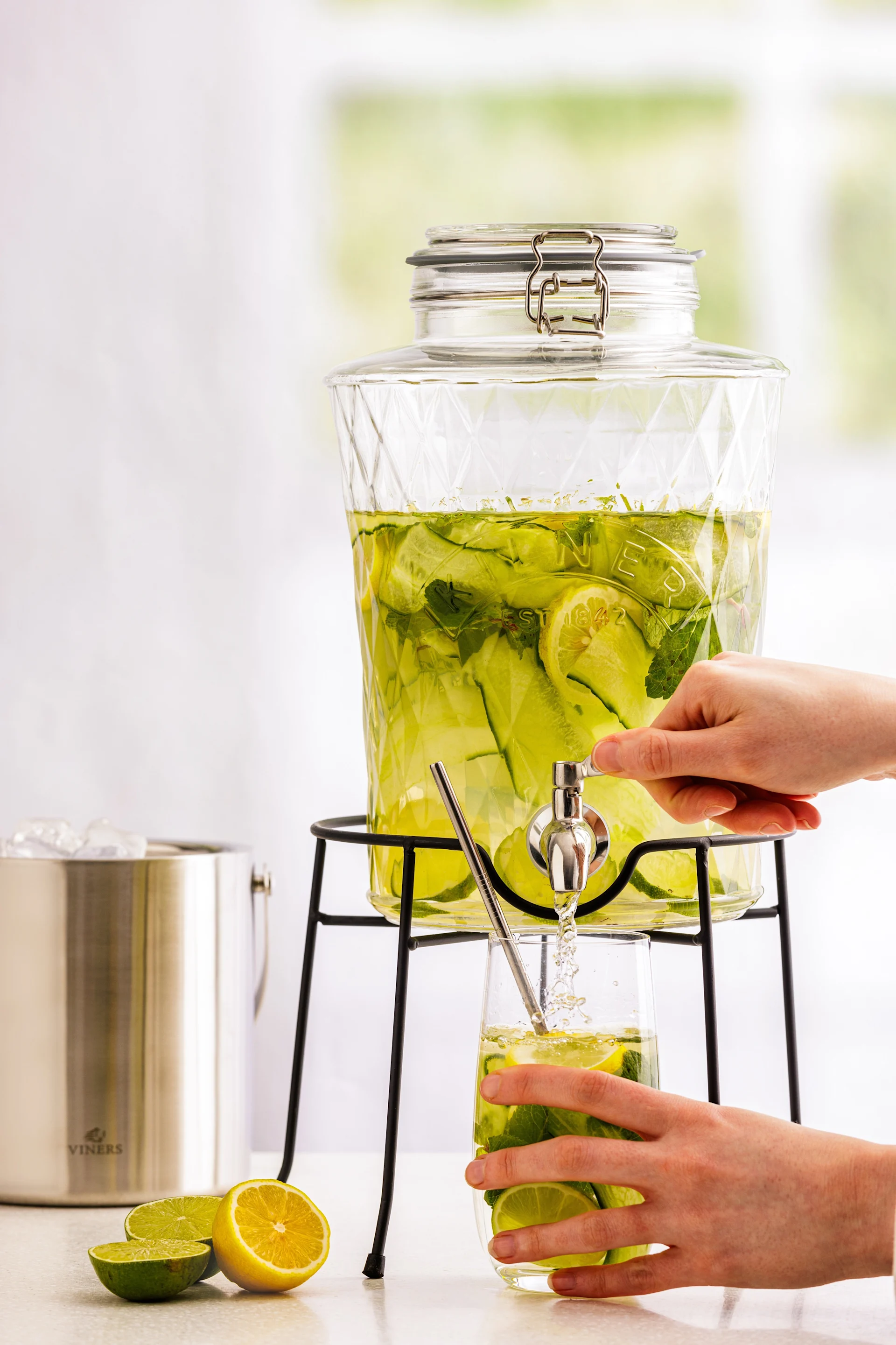 A person's hands fill a glass with refreshing cucumber, lemon, and mint infused water from a clear glass dispenser. A Viners ice bucket and citrus halves are nearby.
