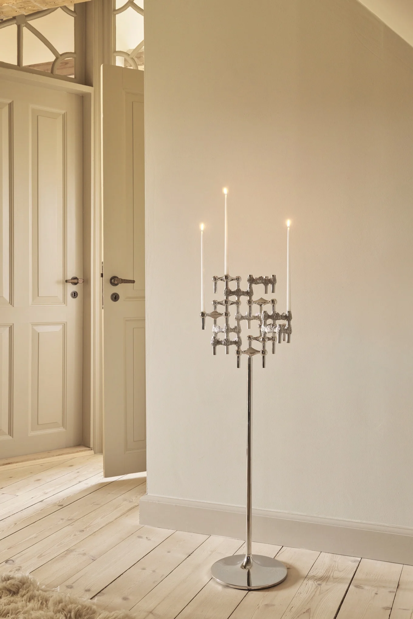 Tall, modern silver candelabra with lit white candles standing on a wooden floor in a room with light walls and open doors.
