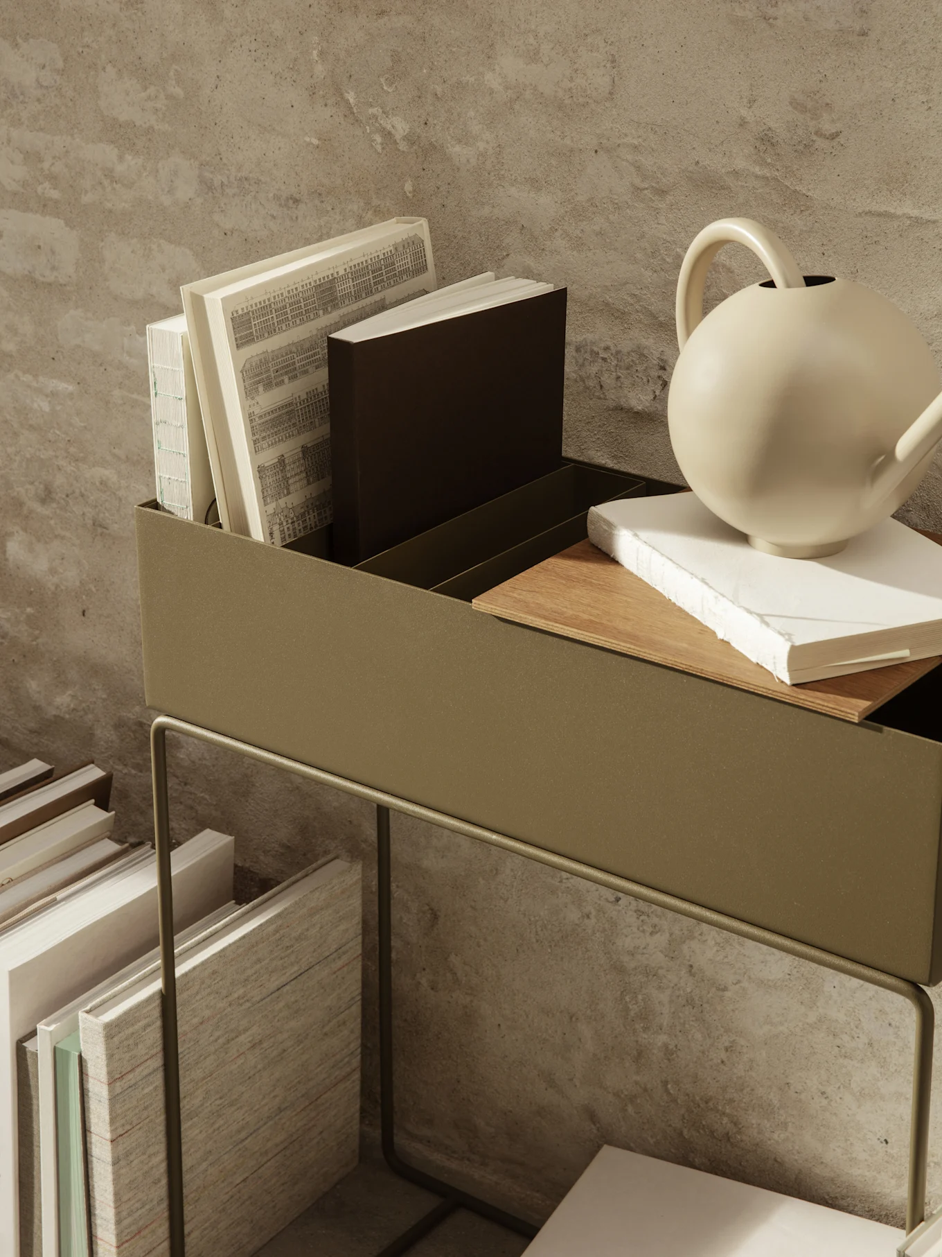 Dark green metal storage unit holding books and a beige watering can, with more books stacked below on the floor.