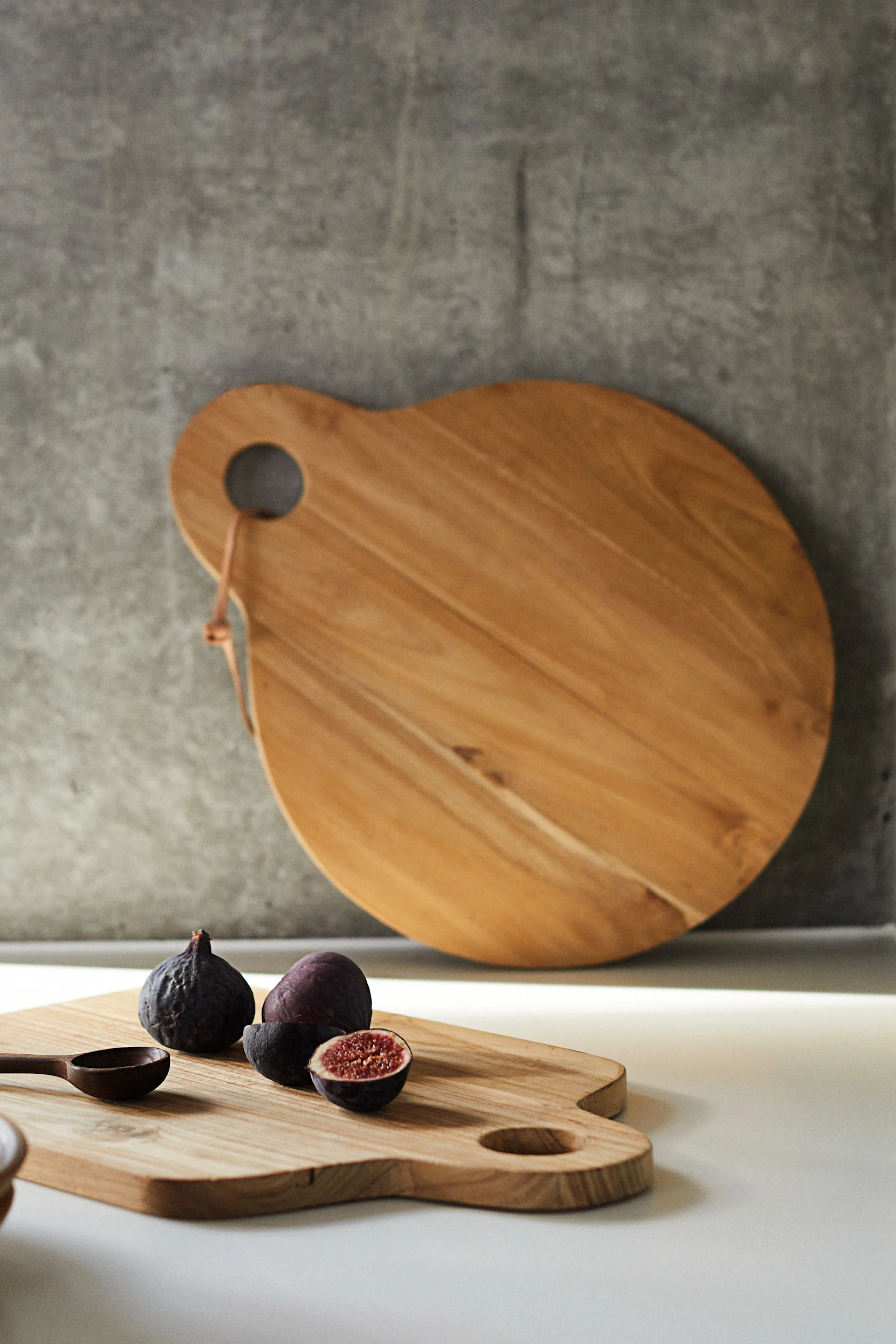 Wooden cutting boards: a large one leans against a gray wall, a smaller one holds figs and a wooden spoon on a white counter.