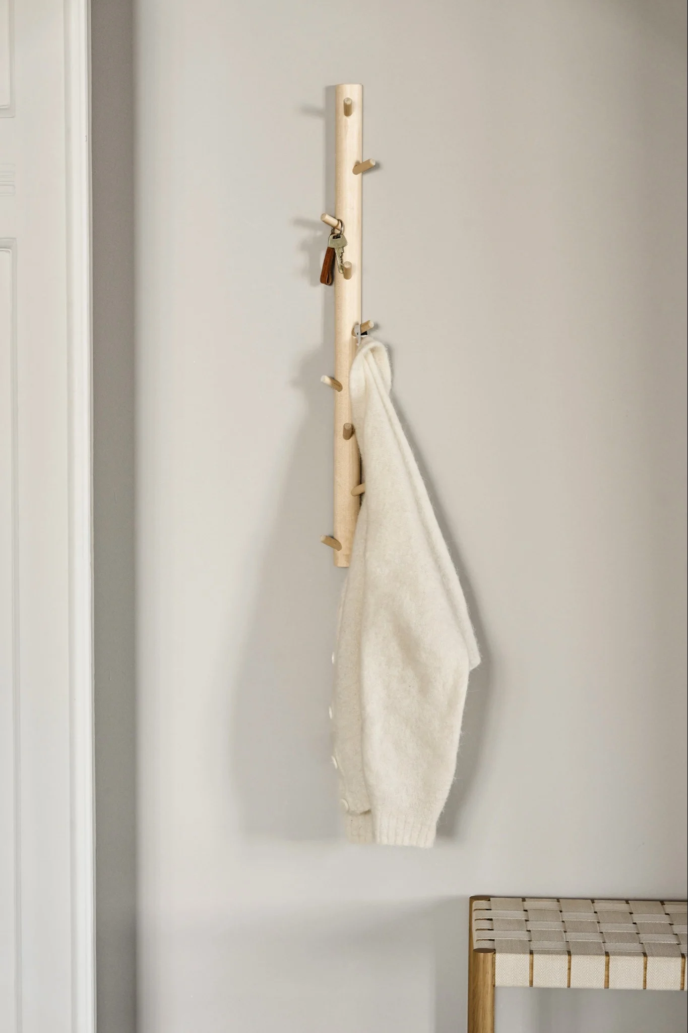 A light wooden coat rack with pegs holds keys and a cream sweater, with a woven bench below.