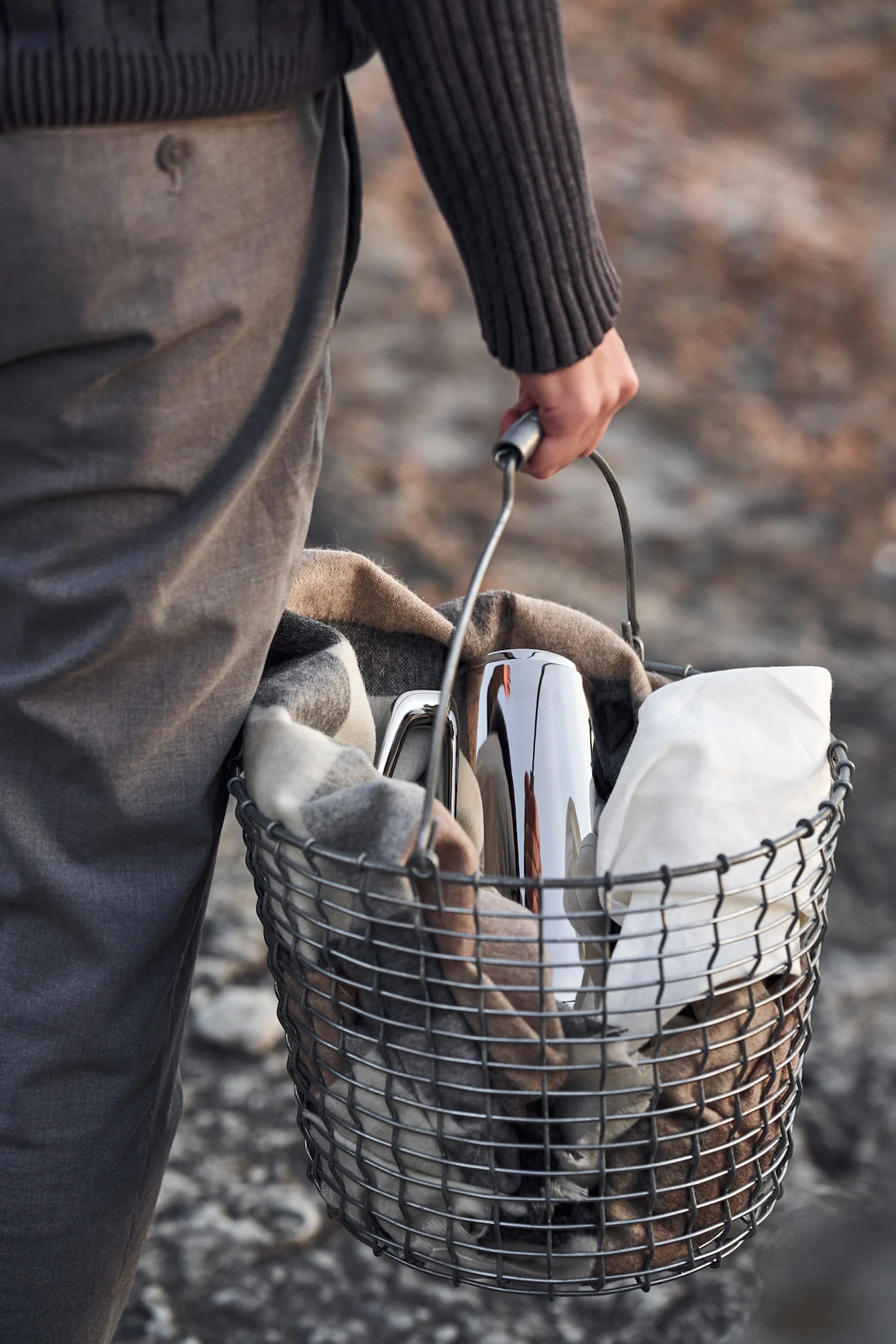 Person carrying a metal basket with a plaid blanket, a silver thermal carafe, and a white cloth.