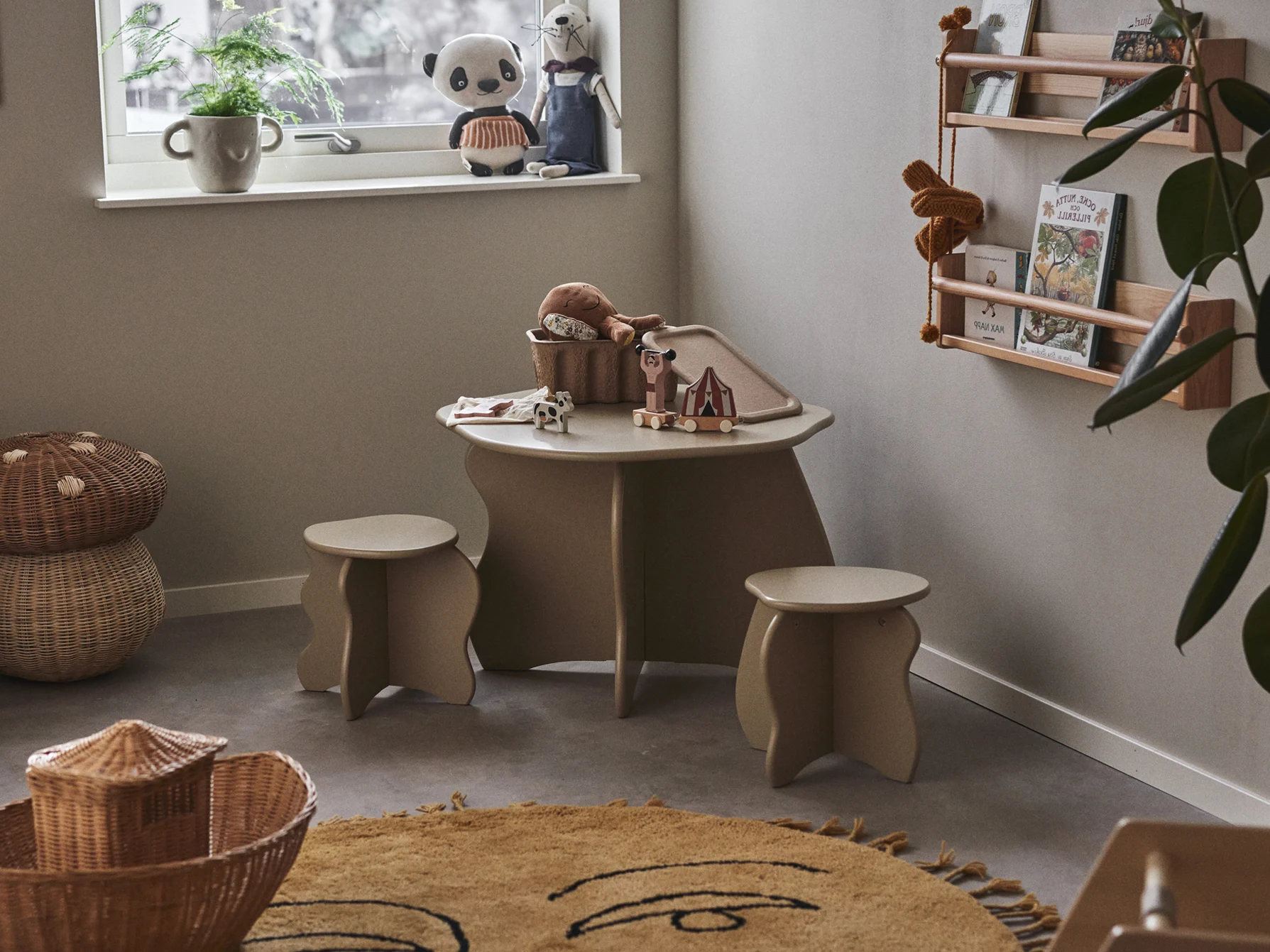 Children's play area with a light brown table, two stools, various toys, woven baskets, and a yellow rug with a face design.