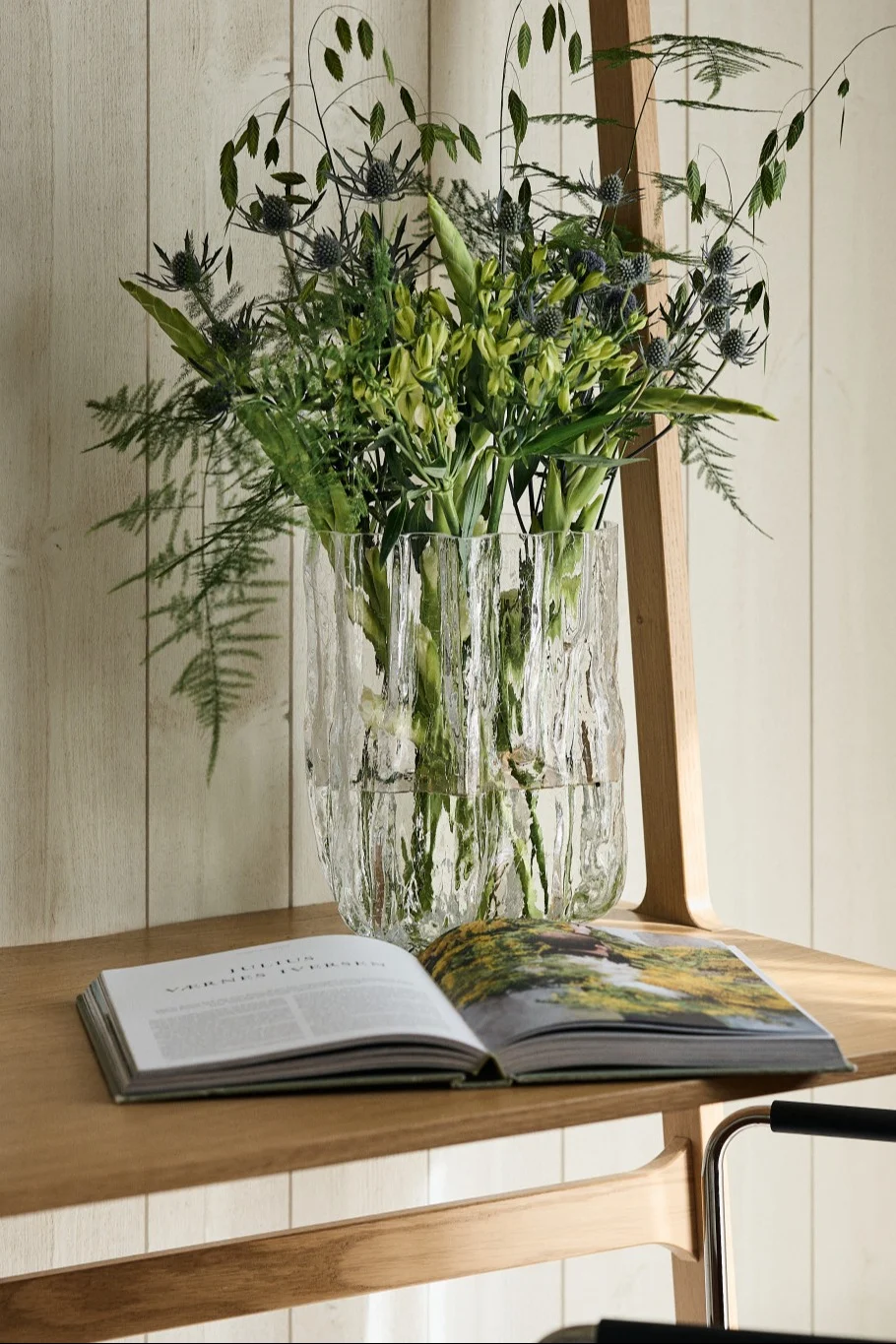 Textured glass vase filled with a bouquet of green thistles and foliage, next to an open book on a light wooden table.
