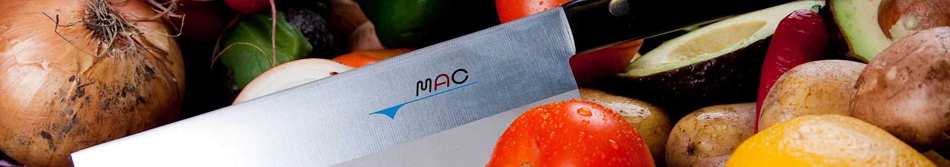 A MAC kitchen knife with its blade angled across a vibrant assortment of fresh vegetables including onions, tomatoes, and avocados.