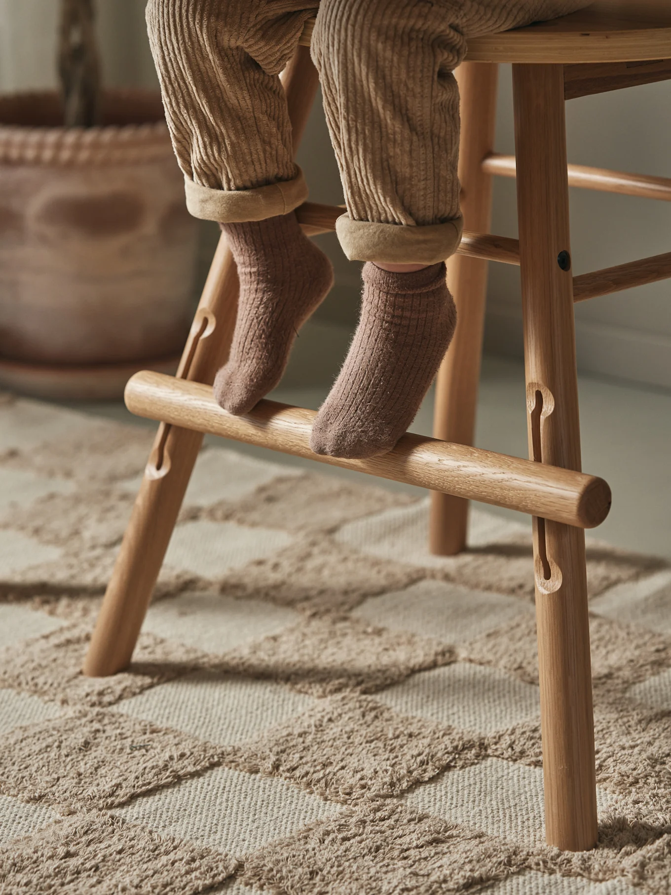 Child's legs in brown corduroy trousers and socks, resting on a wooden high chair footrest on a textured rug.