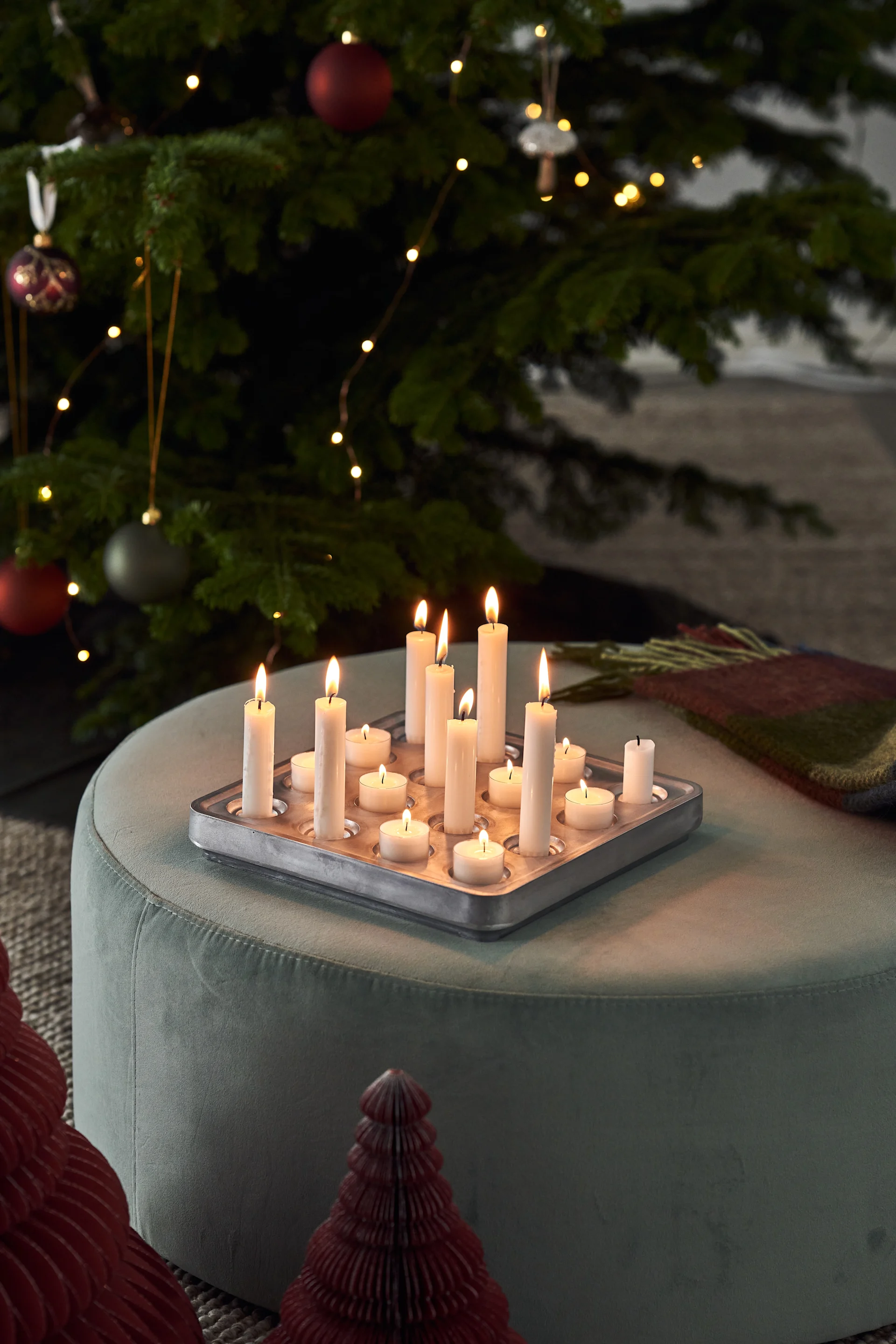 Lit pillar and tea light candles in a metal tray on a green ottoman, with a decorated Christmas tree.