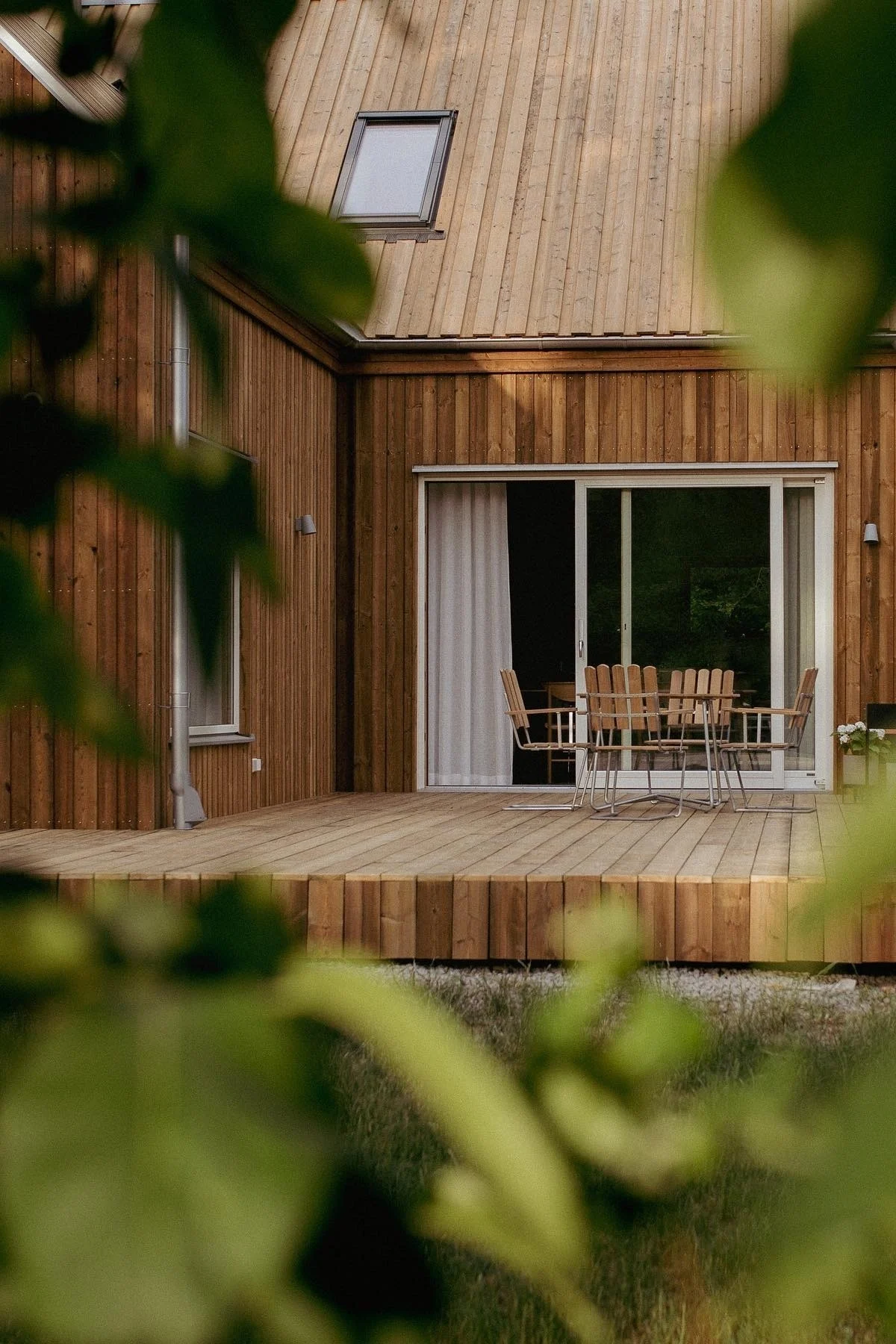 Wooden house with a modern deck, outdoor dining set, and a large sliding glass door, surrounded by greenery.