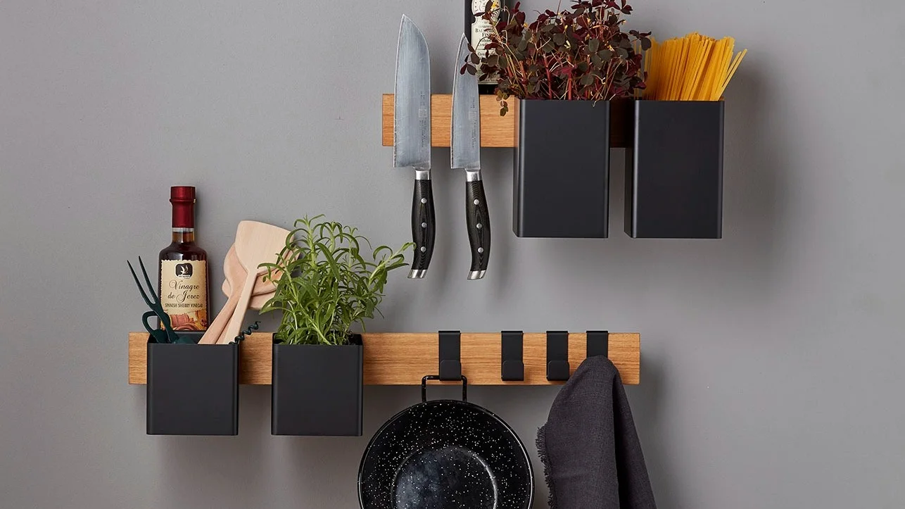 A wall-mounted kitchen organizer featuring wooden shelves and black containers with knives, herbs, pasta, vinegar, pan, and towel.