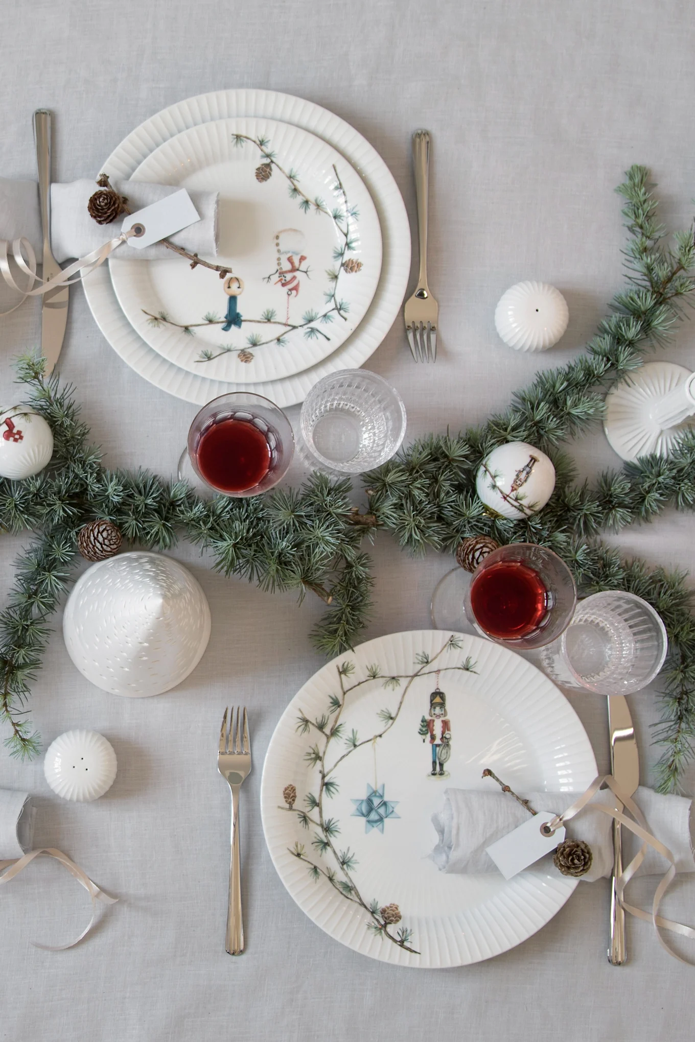 Elegant Christmas table setting for two with white ribbed plates, pine garland, red drinks, and silver cutlery on a light tablecloth.