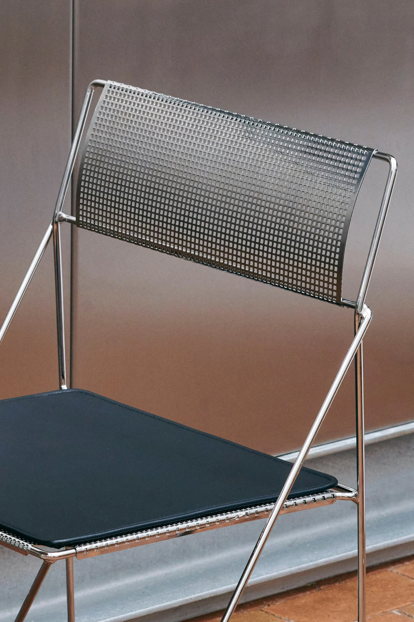 Close-up of a modern folding chair with a chrome frame, a perforated metal backrest, and a dark upholstered seat.