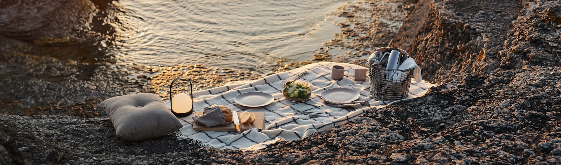 Picnic set up on rocky shore by golden-hour sea with blanket, bread, salad, cups, lantern, cushion, and a basket.
