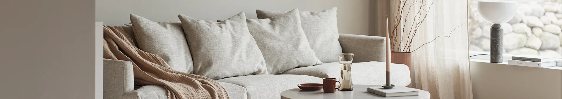 Light sofa with beige throw blanket, coffee table holding a candle and drinkware, and a modern lamp by a window.