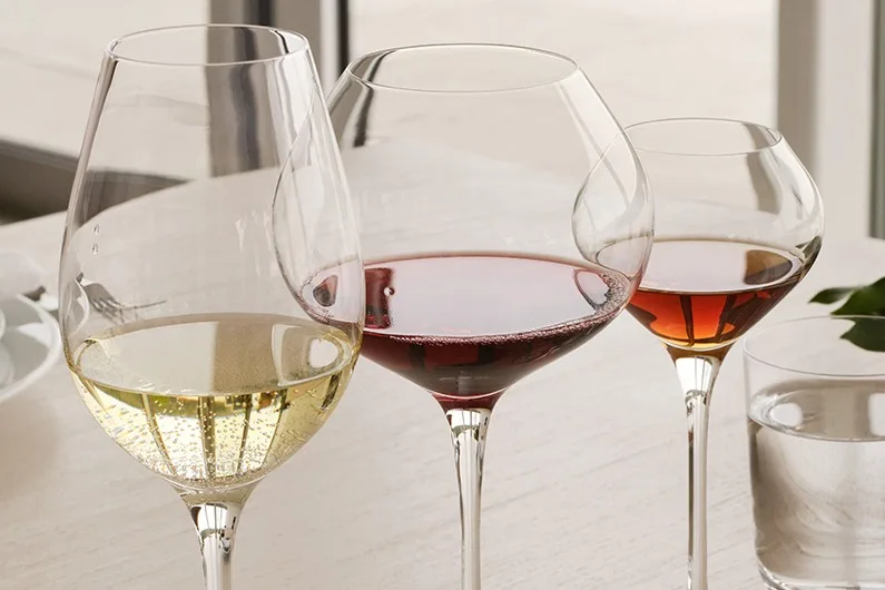 Three wine glasses, one with white wine, one with red, and one with amber wine, with a glass of water on a light table.