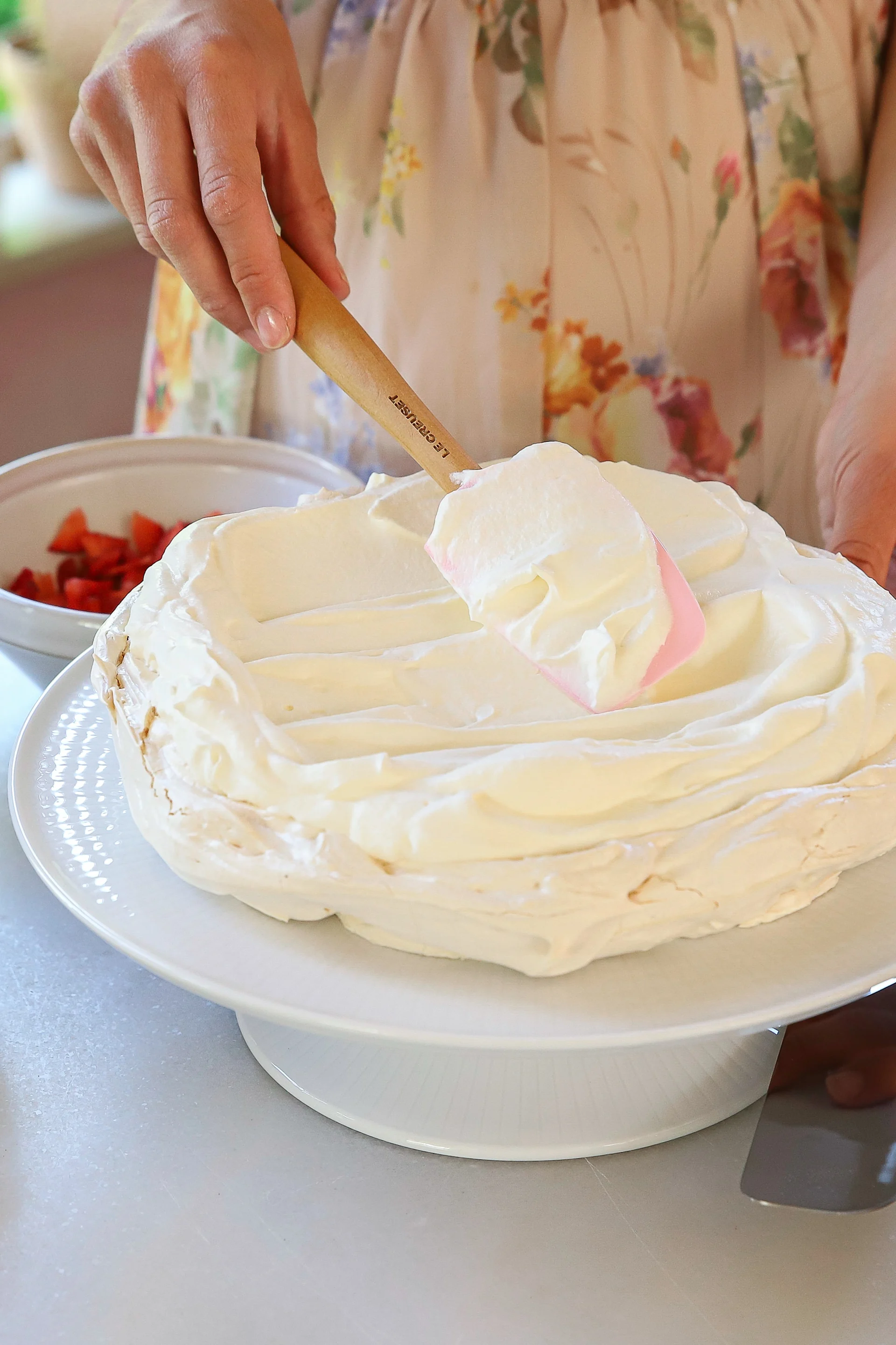 Hands spreading whipped cream on a pavlova with a Le Creuset spatula; bowl of strawberries in background.
