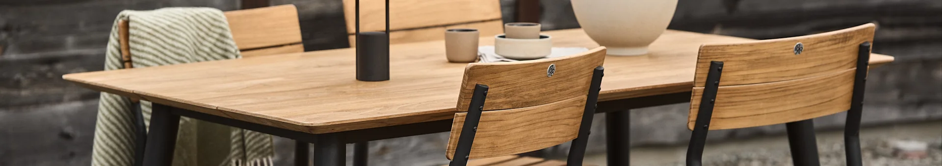 Outdoor dining set with a rectangular wooden table, wooden chairs with black frames, ceramic dishes, and a candle holder.