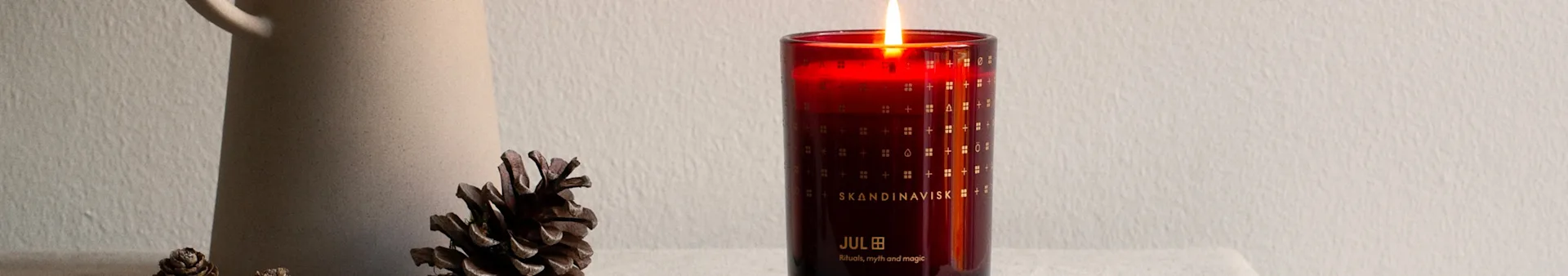 A lit red candle in a glass holder with 'Skandinavisk Jul' text, next to a ceramic pitcher and pinecones.