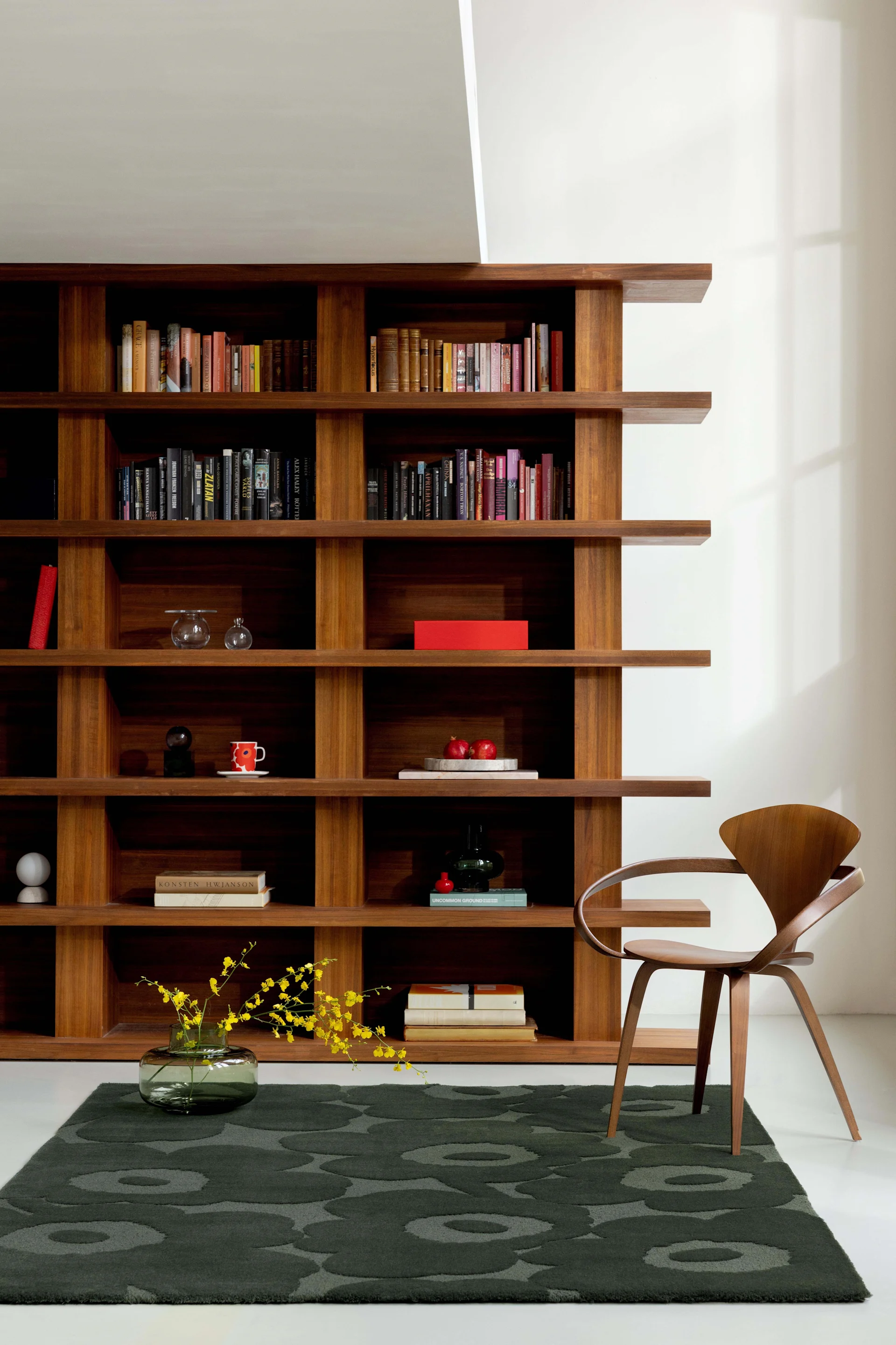 A stylish room featuring a tall wooden bookshelf filled with books and decor, a curved wooden chair, and a dark green rug.