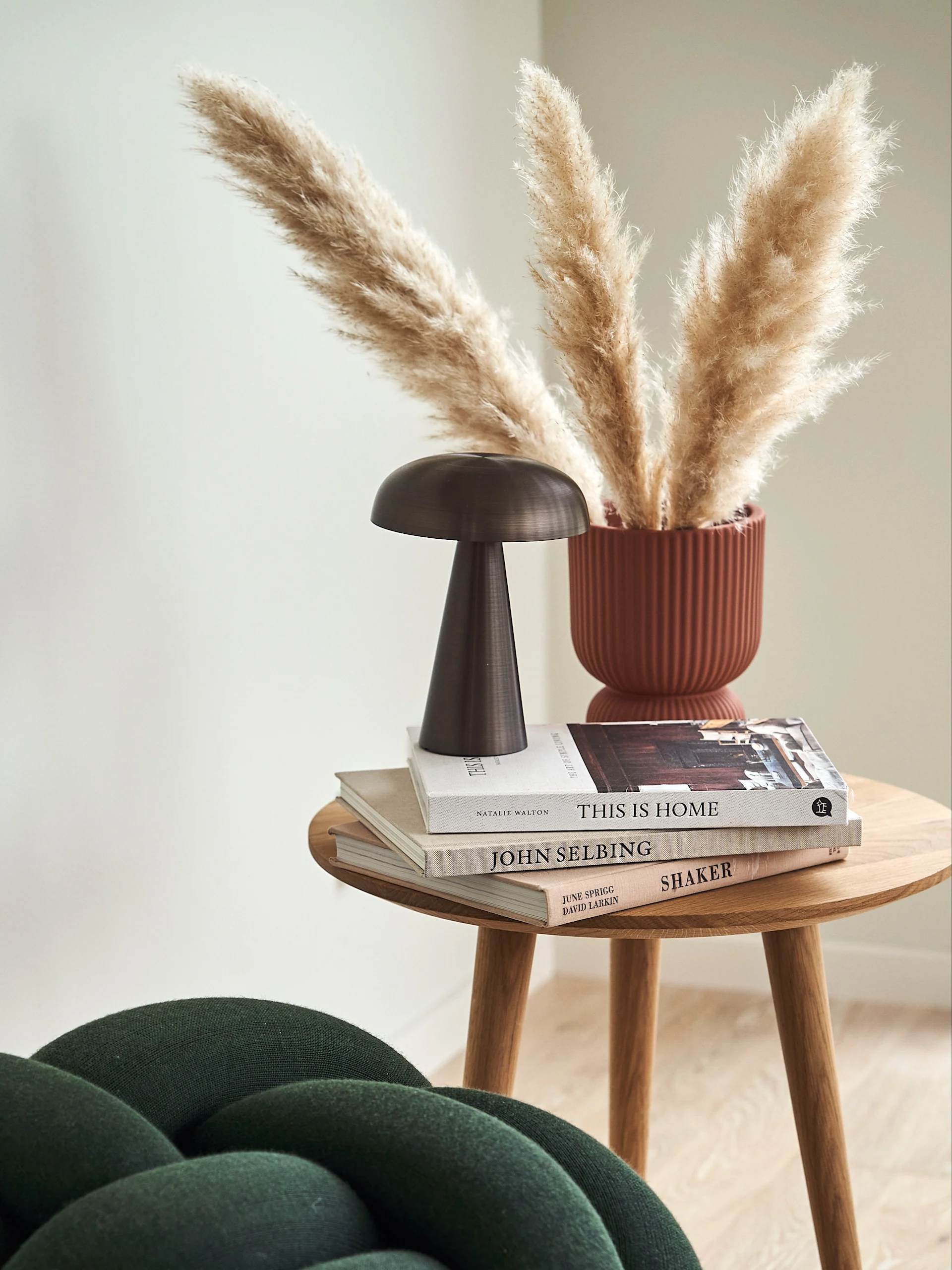 Round wooden side table with stacked books, a dark bronze mushroom lamp, and a rust-colored vase holding pampas grass.