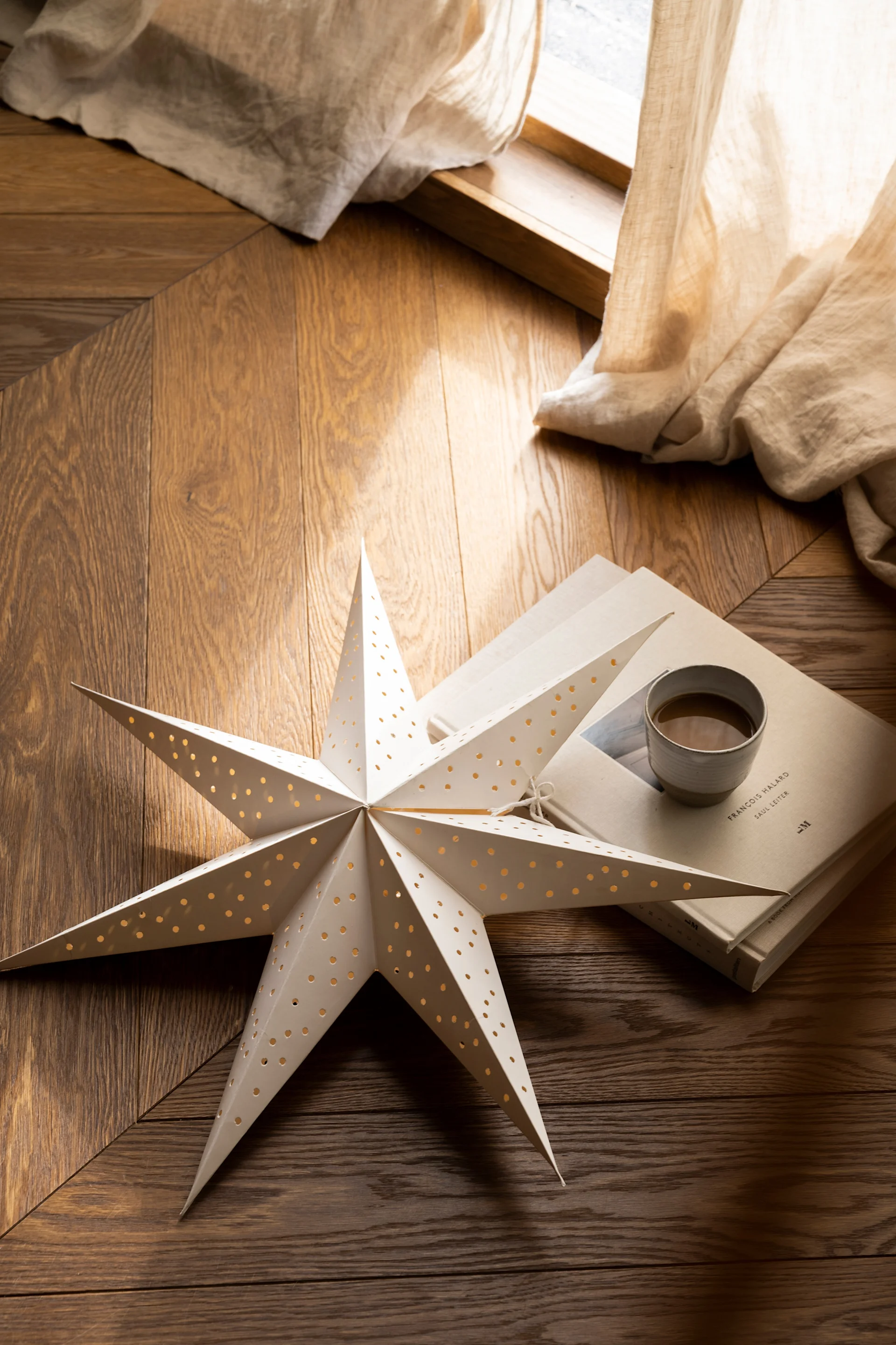 An illuminated white paper star with cutouts lies on a wooden floor next to two books and a ceramic cup, with a light curtain in the background.