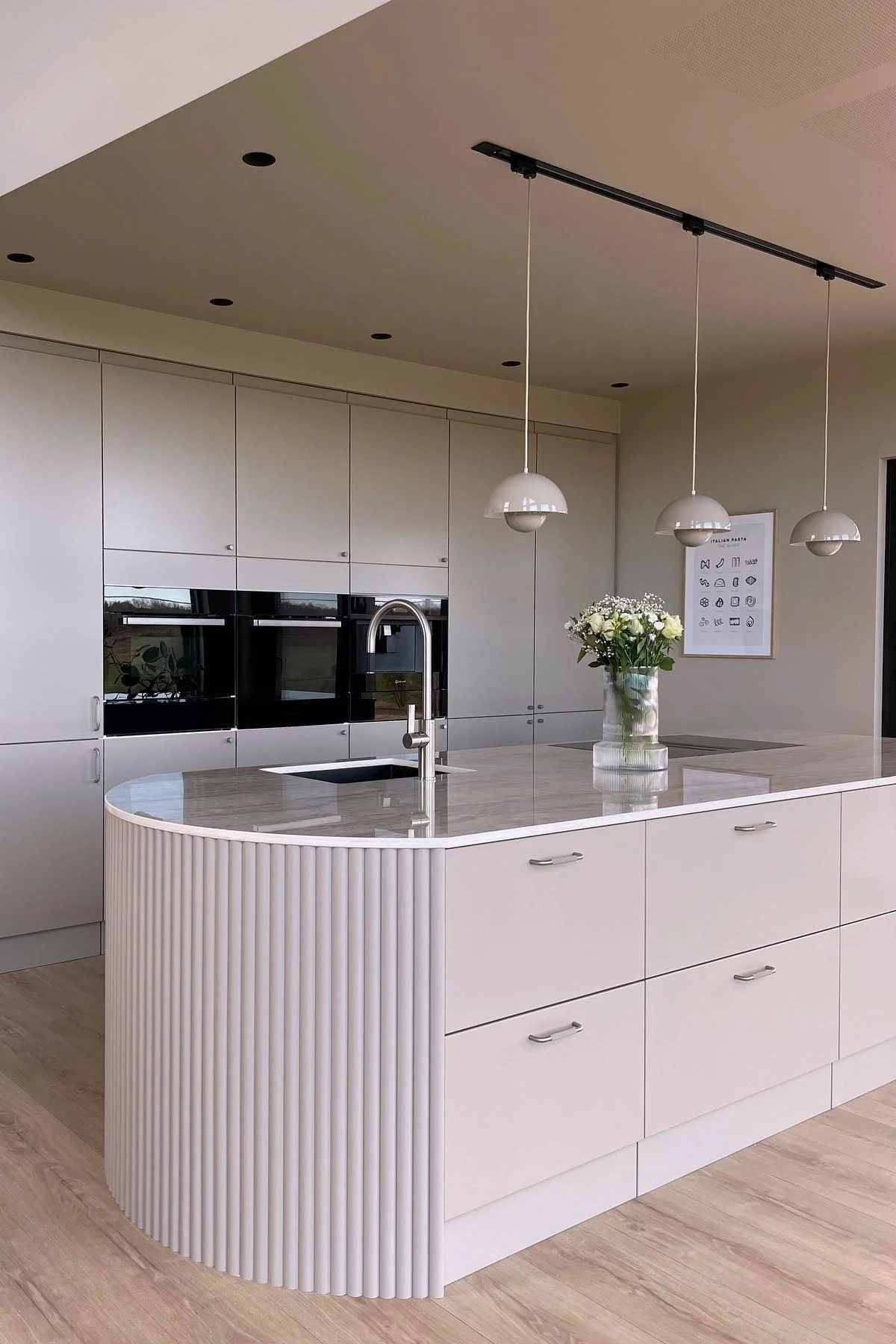 Modern kitchen with light cabinetry, a curved island featuring a sink, stainless faucet, pendant lights, and white flowers.
