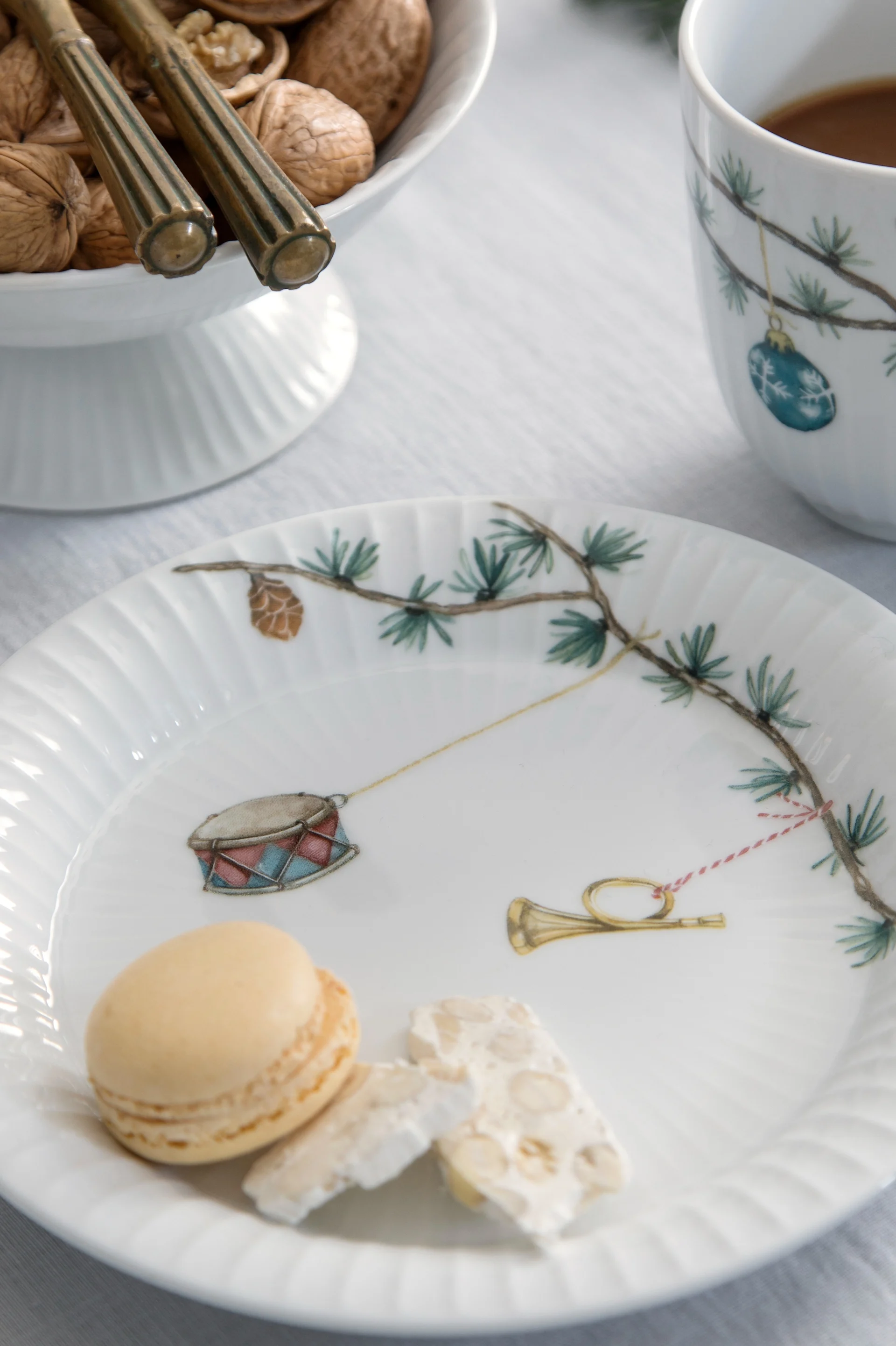 White plate with Christmas decorations holding a macaron and nougat, with walnuts and a mug in the background.