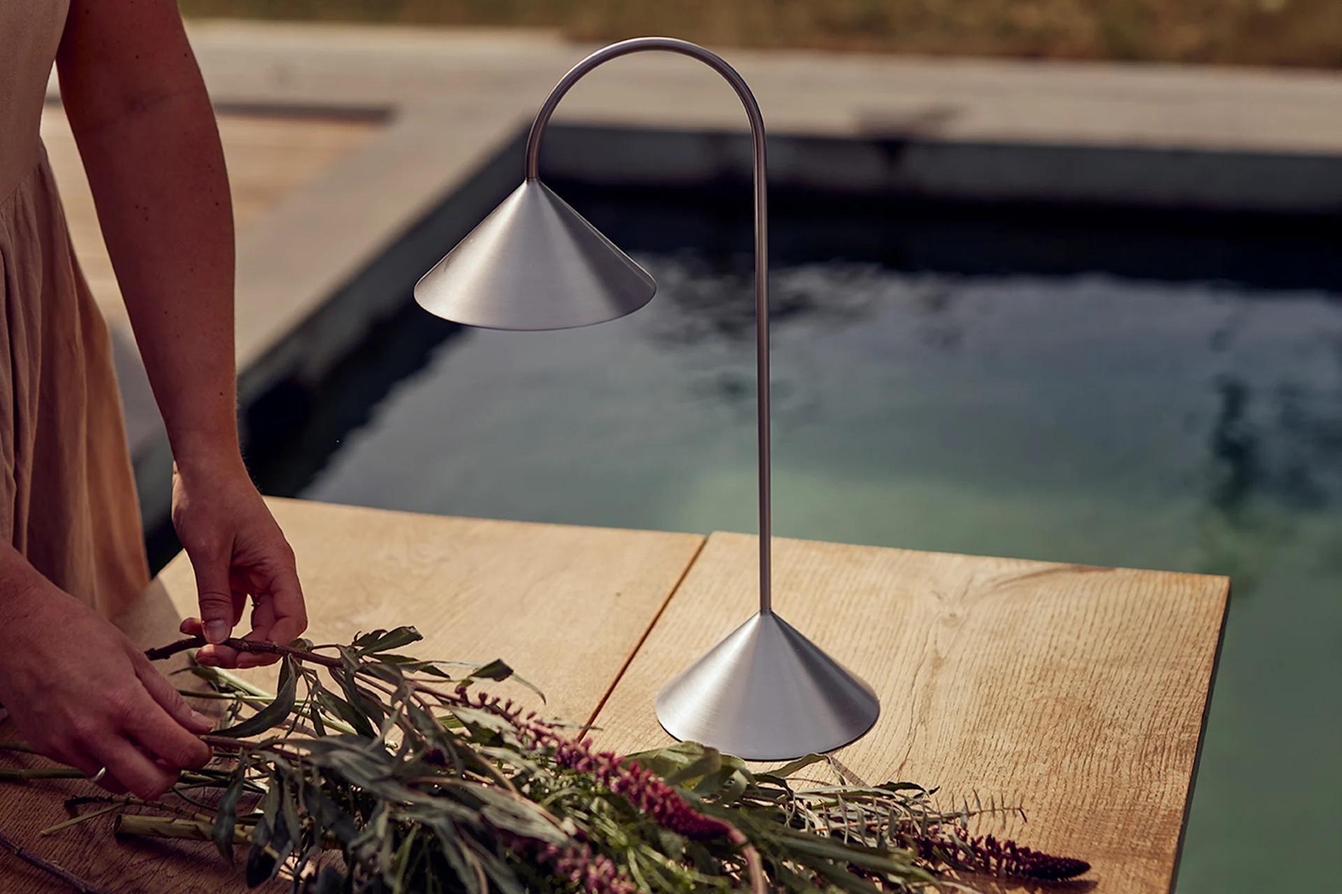 Hands arranging flowers on a wooden table next to a brushed metal table lamp by a pool.